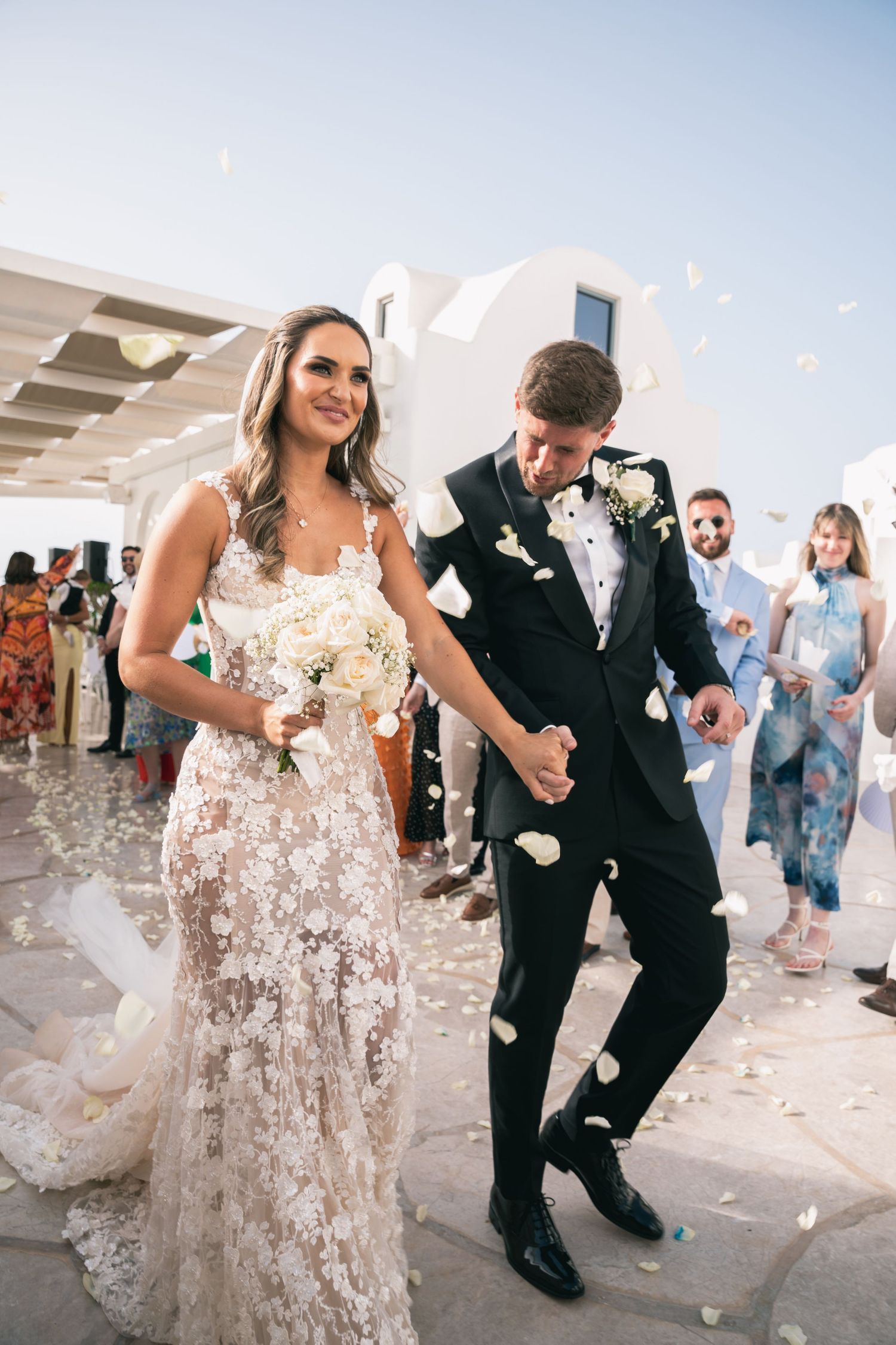 Bride in lace gown and groom in tuxedo walk through rose petal confetti at outdoor wedding ceremony.