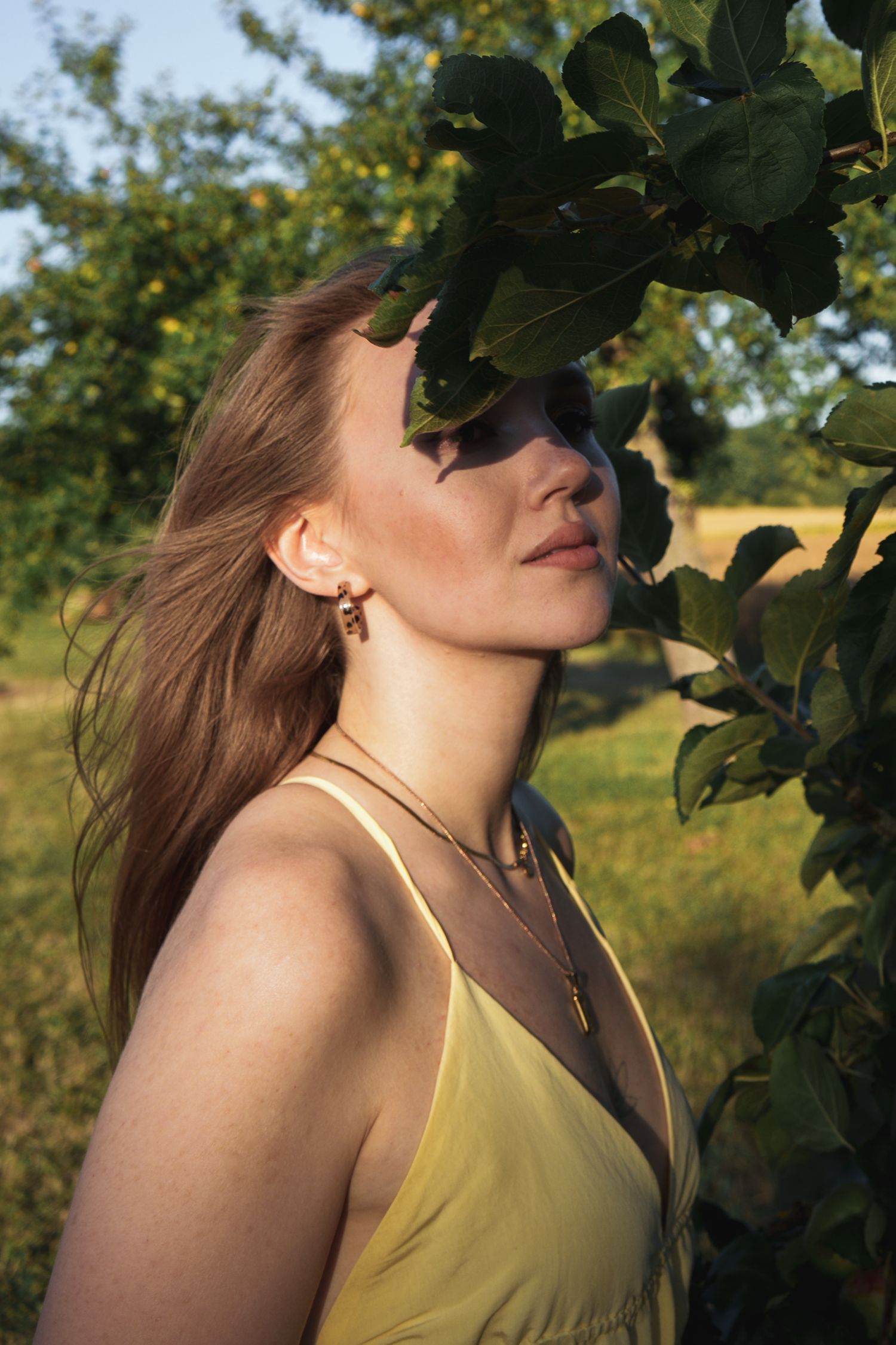 Young woman in yellow dress standing by green leafy tree branches in warm golden sunlight outdoors.