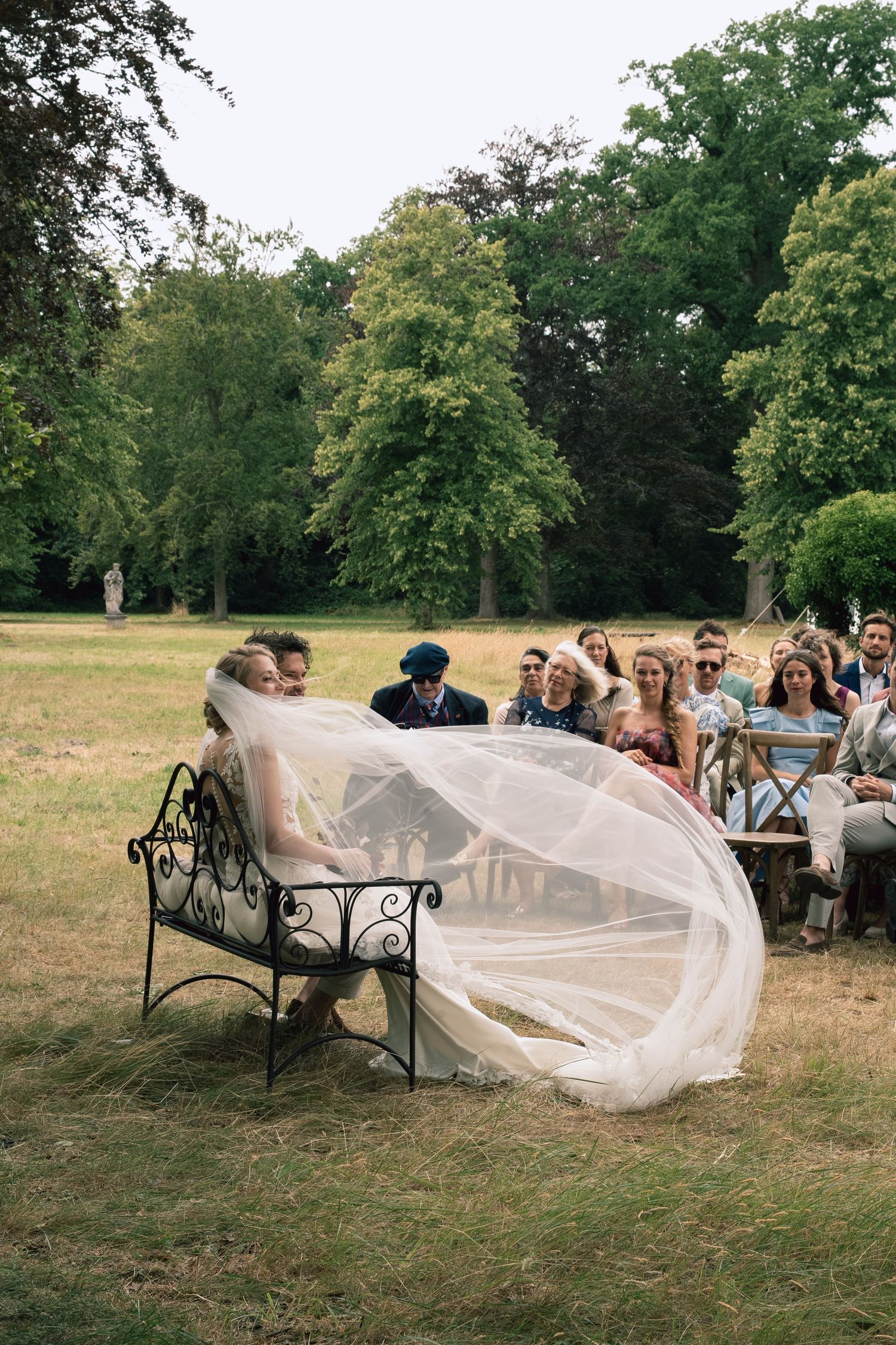 Bride seated on ornate iron bench with flowing veil billowing in wind during outdoor wedding ceremony.