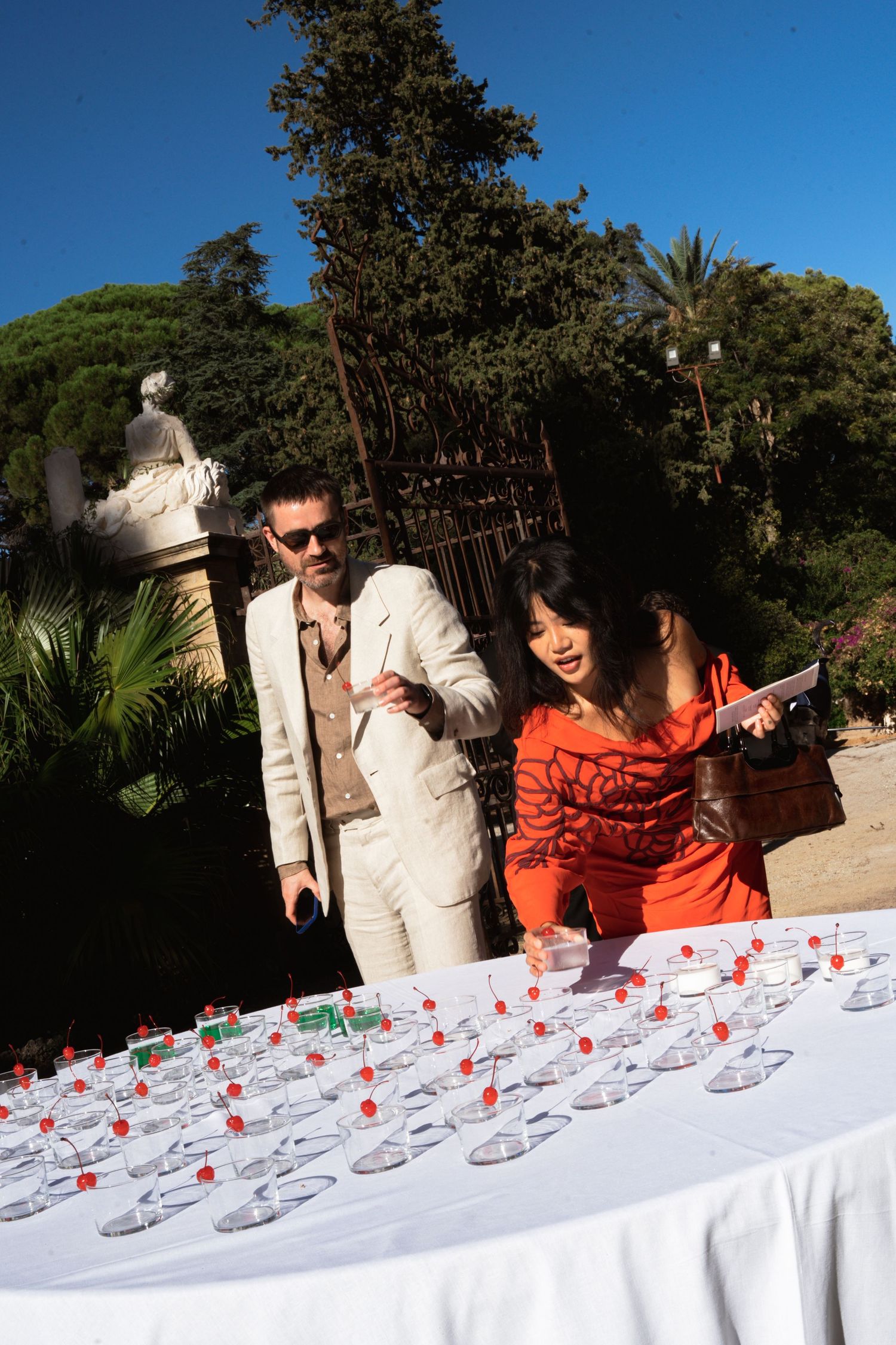 Man in white suit and woman in red outfit browse place cards on a white table at an outdoor event.