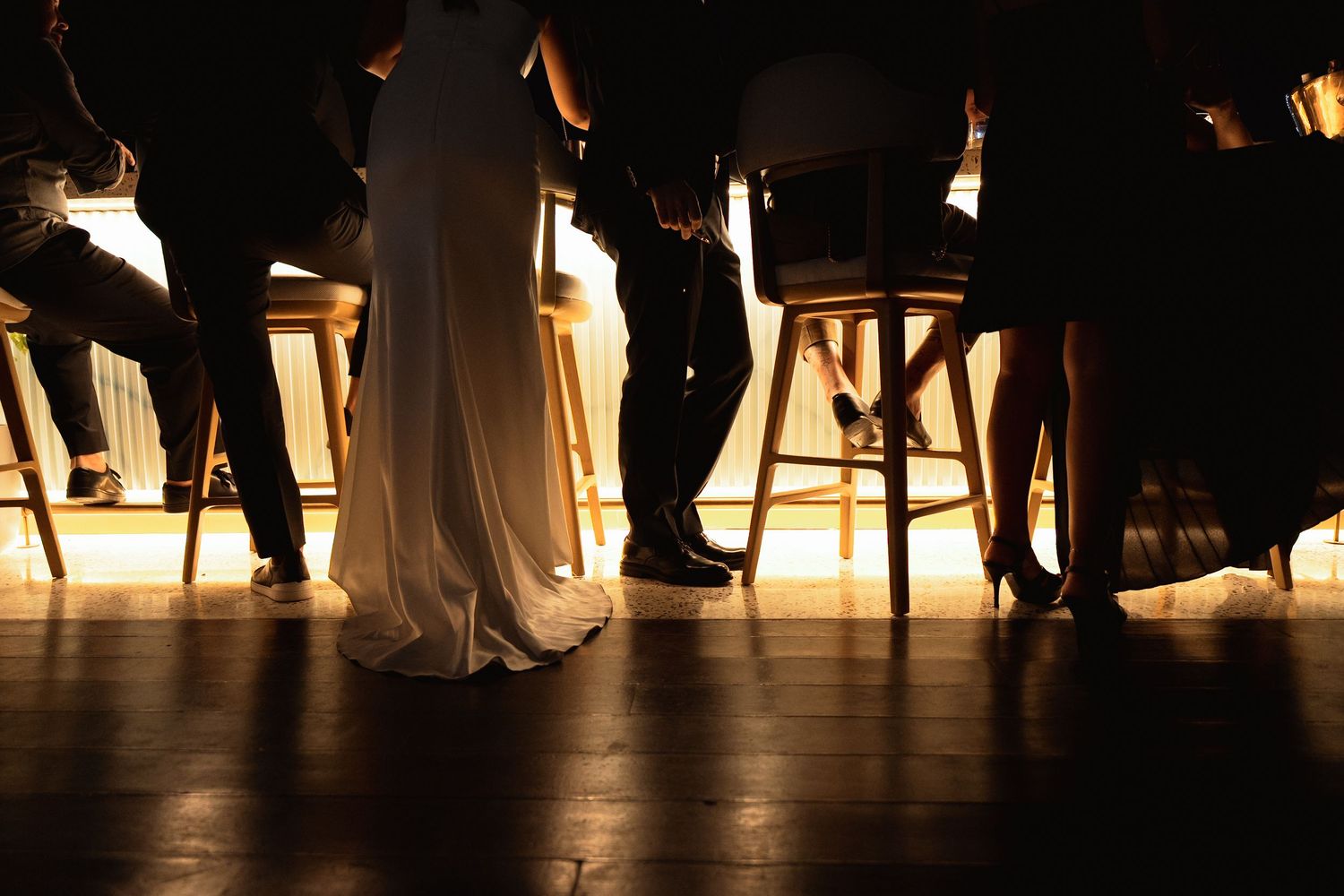 Dramatic low-angle shot of wedding guests and bride in white gown seated at bar stools in warm light.