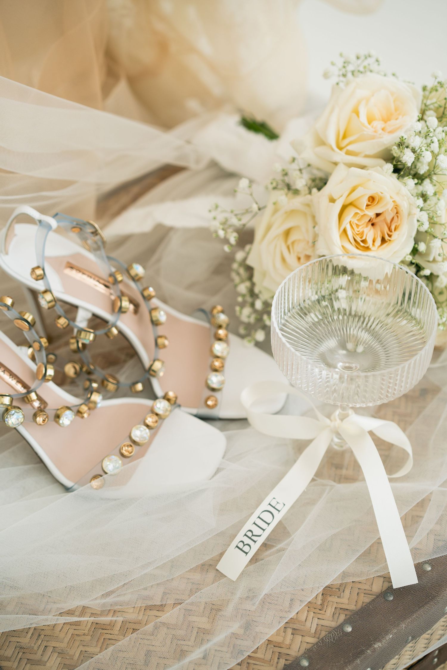 Elegant bridal flat lay with pearl-studded heels, white rose bouquet, crystal glass, and bride ribbon on tulle veil.
