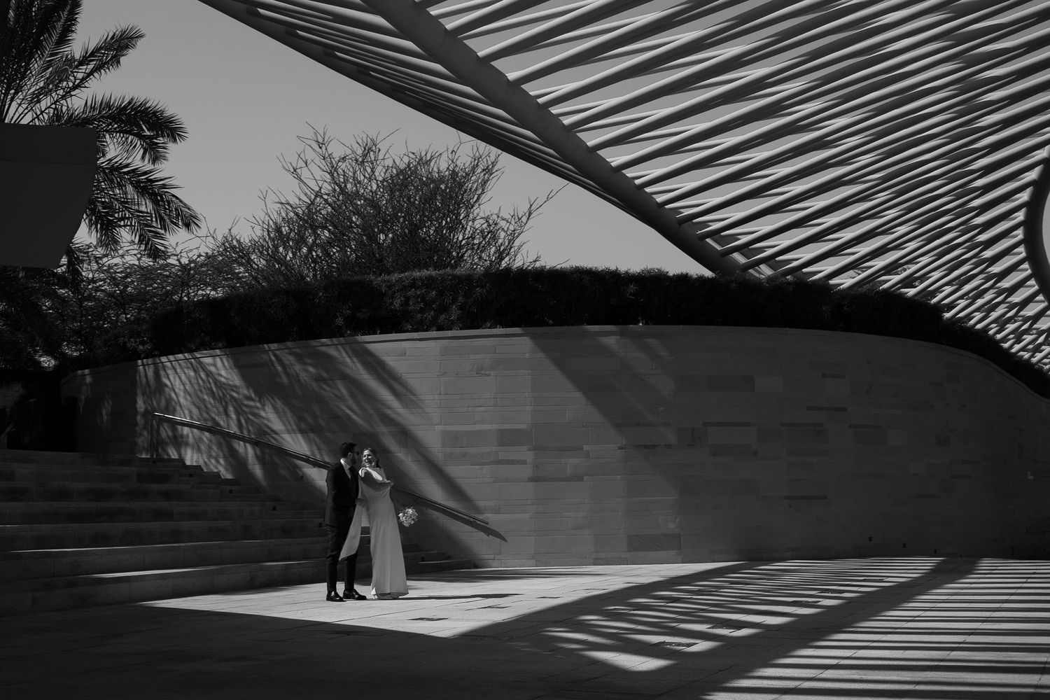 Black and white photo of a couple embracing under a modern architectural structure with dramatic shadow patterns.
