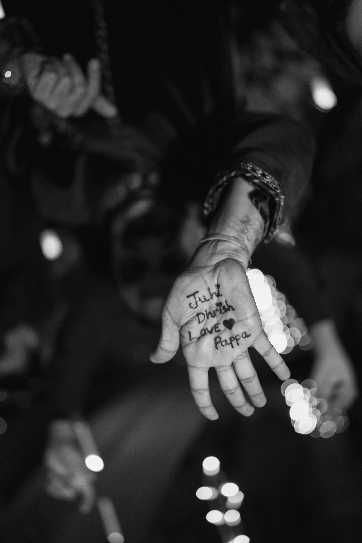 Black and white photo of a hand with writing reading 'Juhi, Dhrish, Love, Pappa' surrounded by bokeh lights.