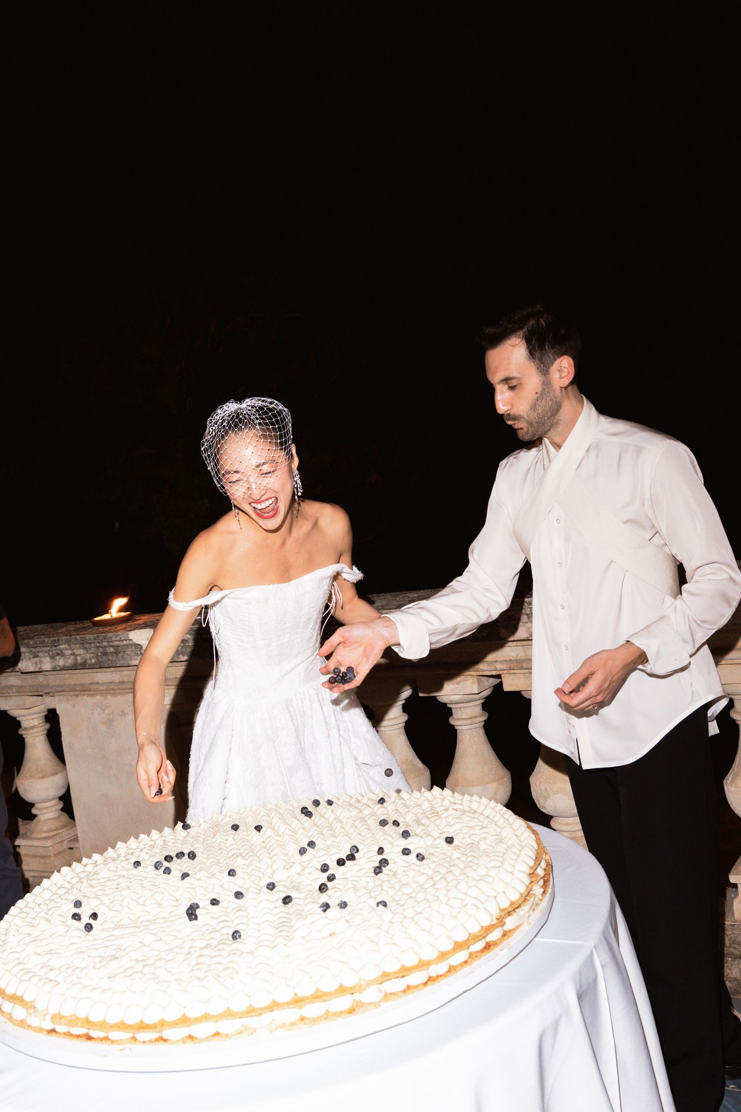 Bride and groom cutting a large white wedding cake decorated with blueberries at an outdoor night reception.