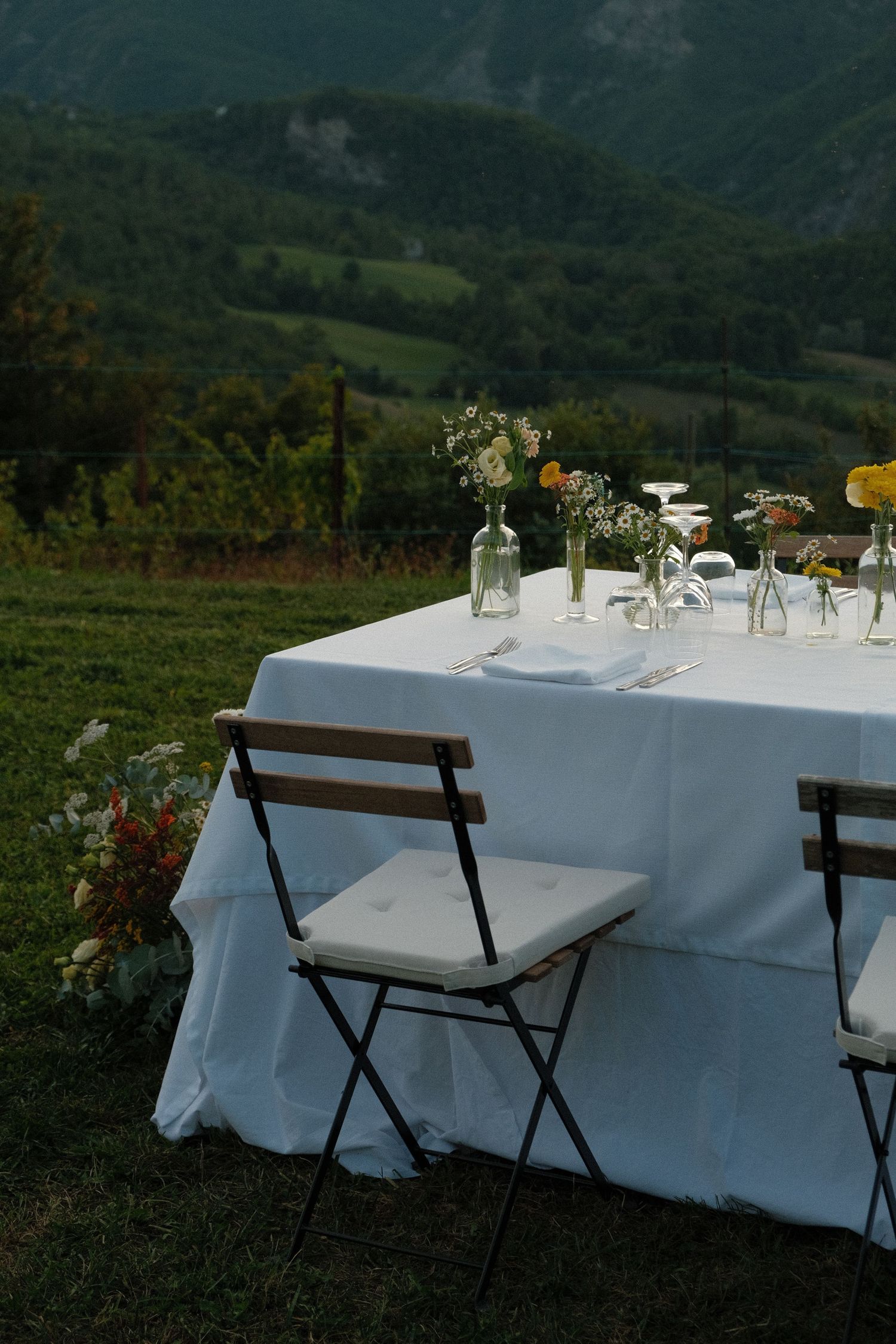 Elegant outdoor dining table with white linen, floral arrangements, and mountain views in a scenic vineyard setting.