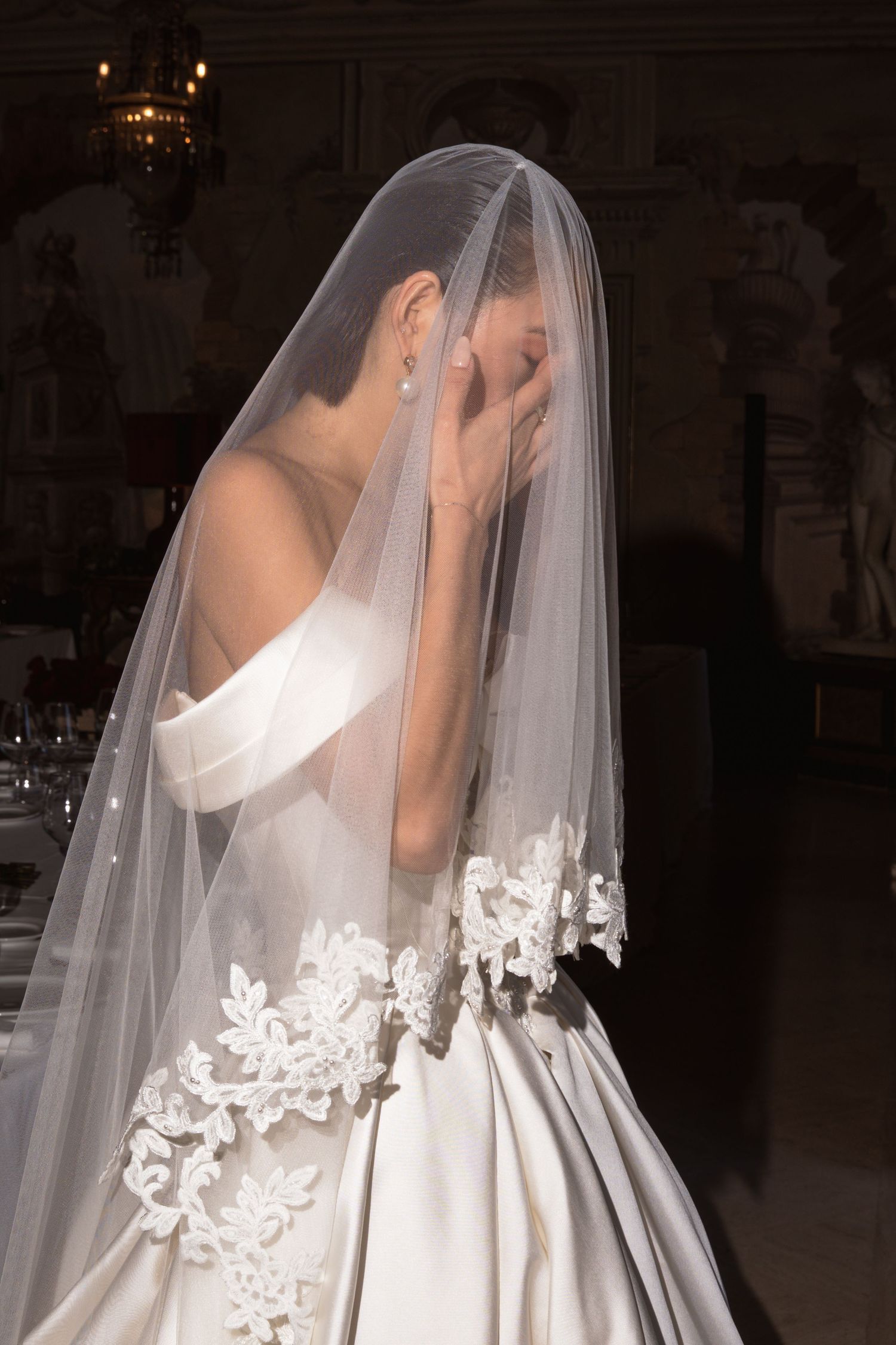 Emotional bride in off-shoulder ball gown with lace-trimmed veil covering her face in dimly lit venue.