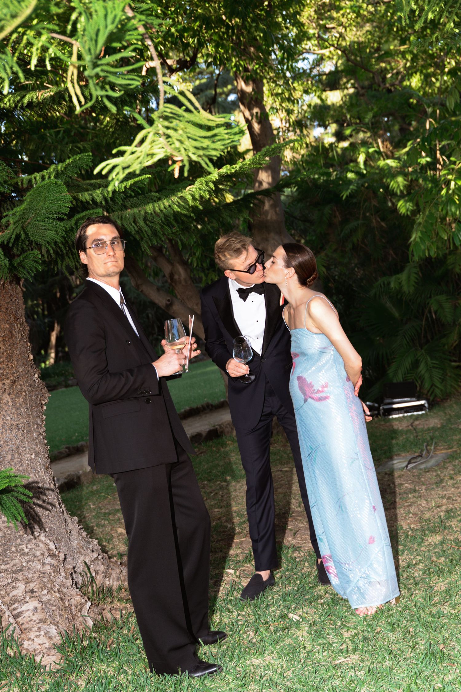 Three elegantly dressed guests at an outdoor evening event, two men in tuxedos and a woman in a blue gown sharing a kiss.
