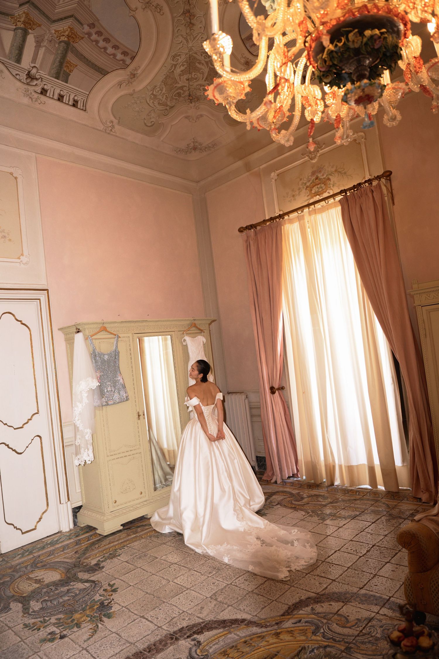 Bride in elegant white ball gown stands in ornate pink room with chandelier and mirror.