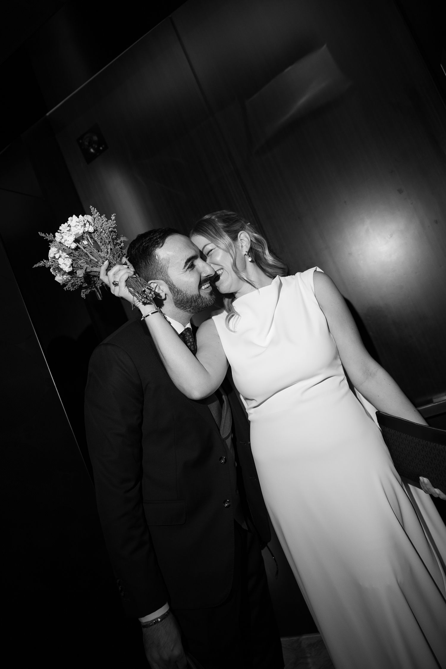 Black and white photo of newlyweds sharing a kiss while bride holds colorful bouquet indoors.