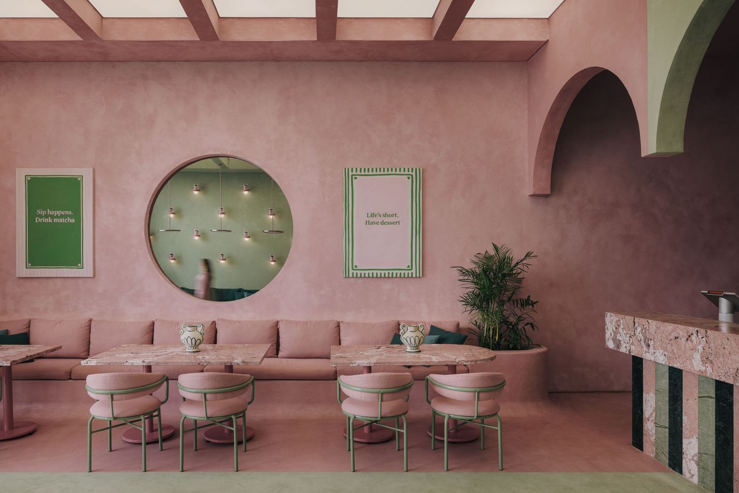 Pink-themed restaurant interior with circular mirror, green framed artwork, long banquette seating, and arched doorways.