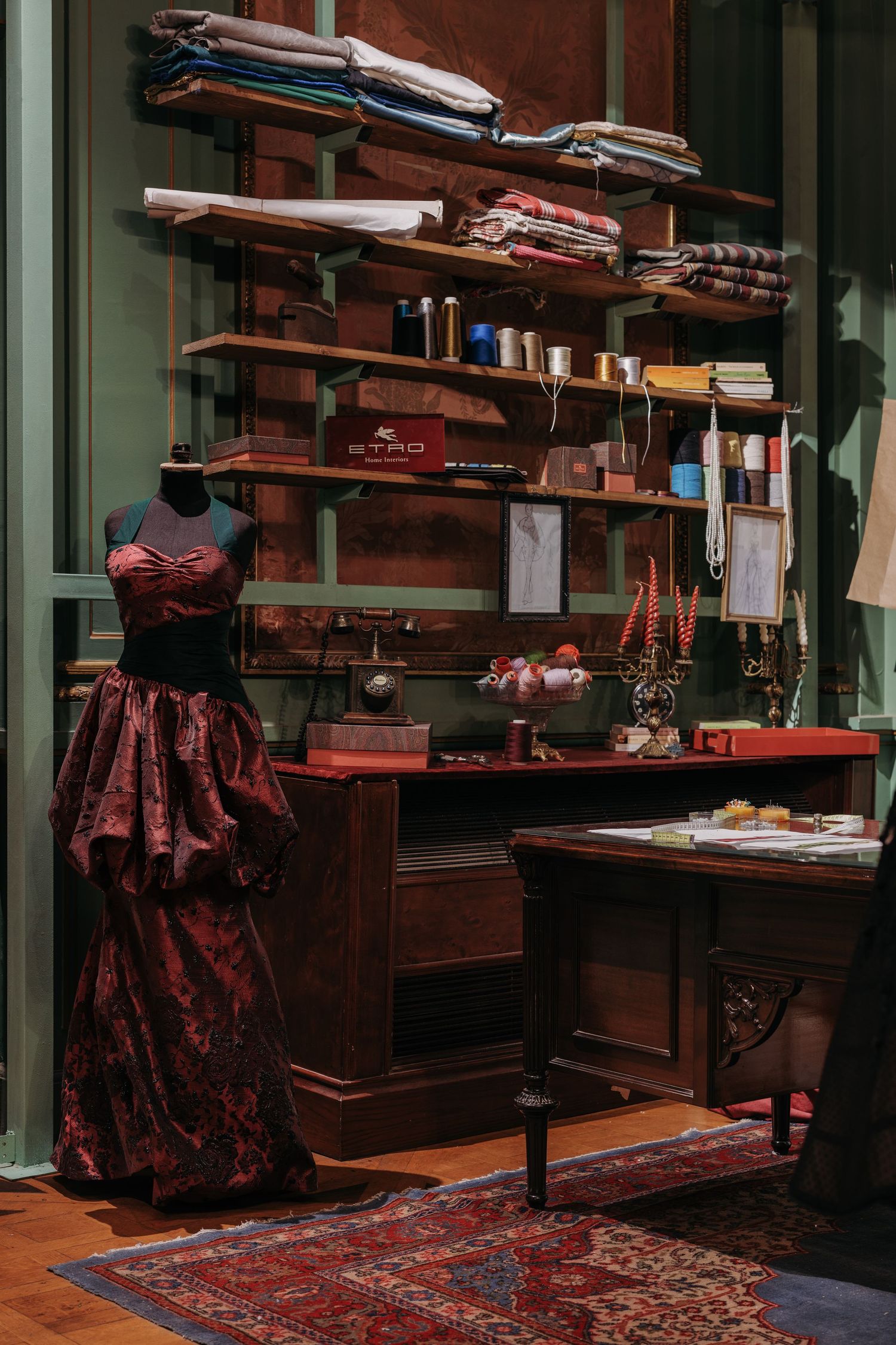Vintage tailor's atelier with a red gown on mannequin, wooden desk, fabric shelves, and sewing supplies.