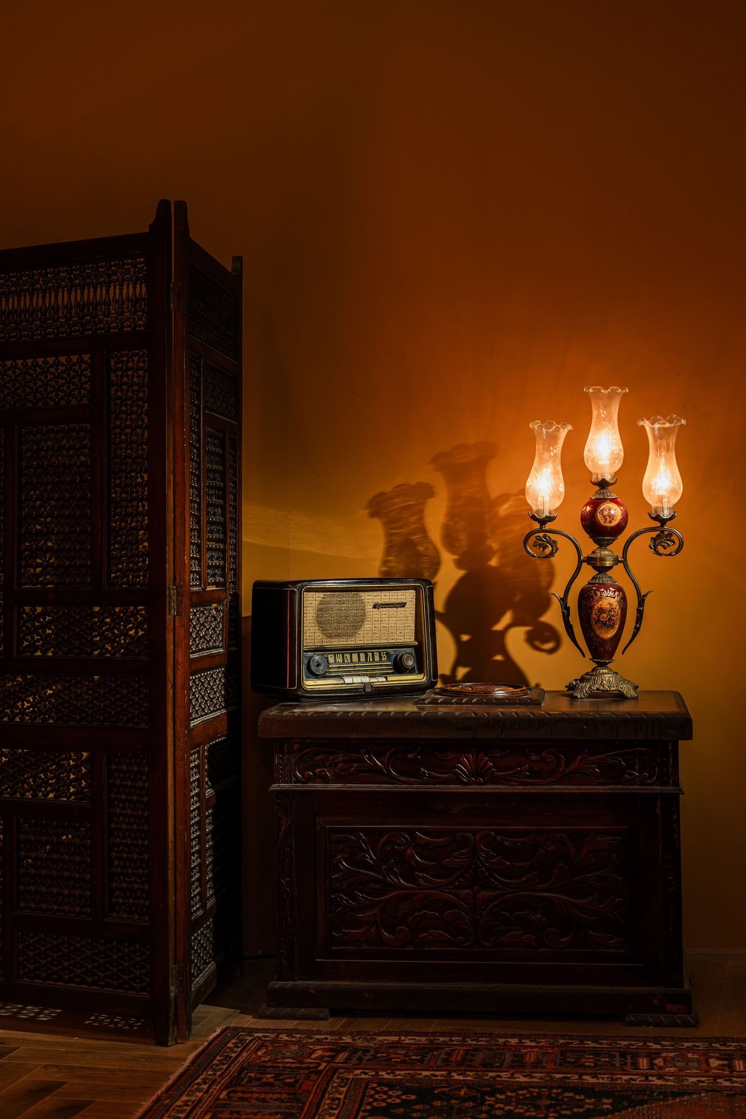 Vintage radio and ornate candelabra lamp on carved wooden chest beside decorative folding screen.