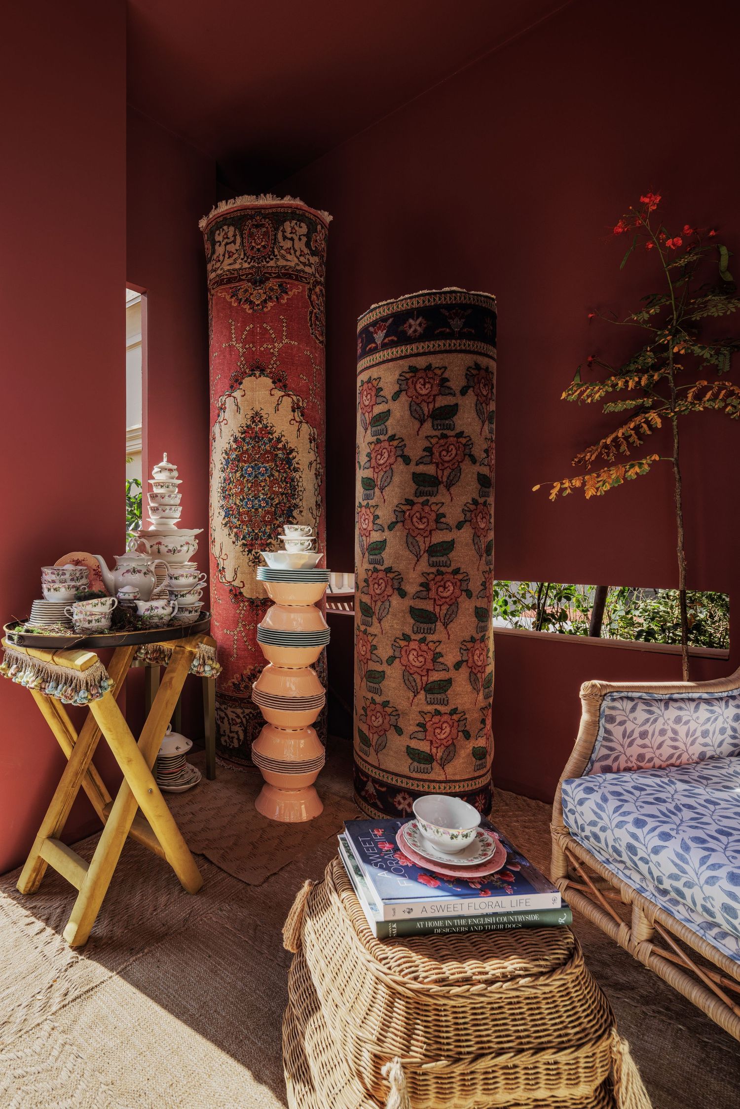 Crimson room with rolled decorative carpets, wicker furniture, ceramic collection, and lush plant accents.