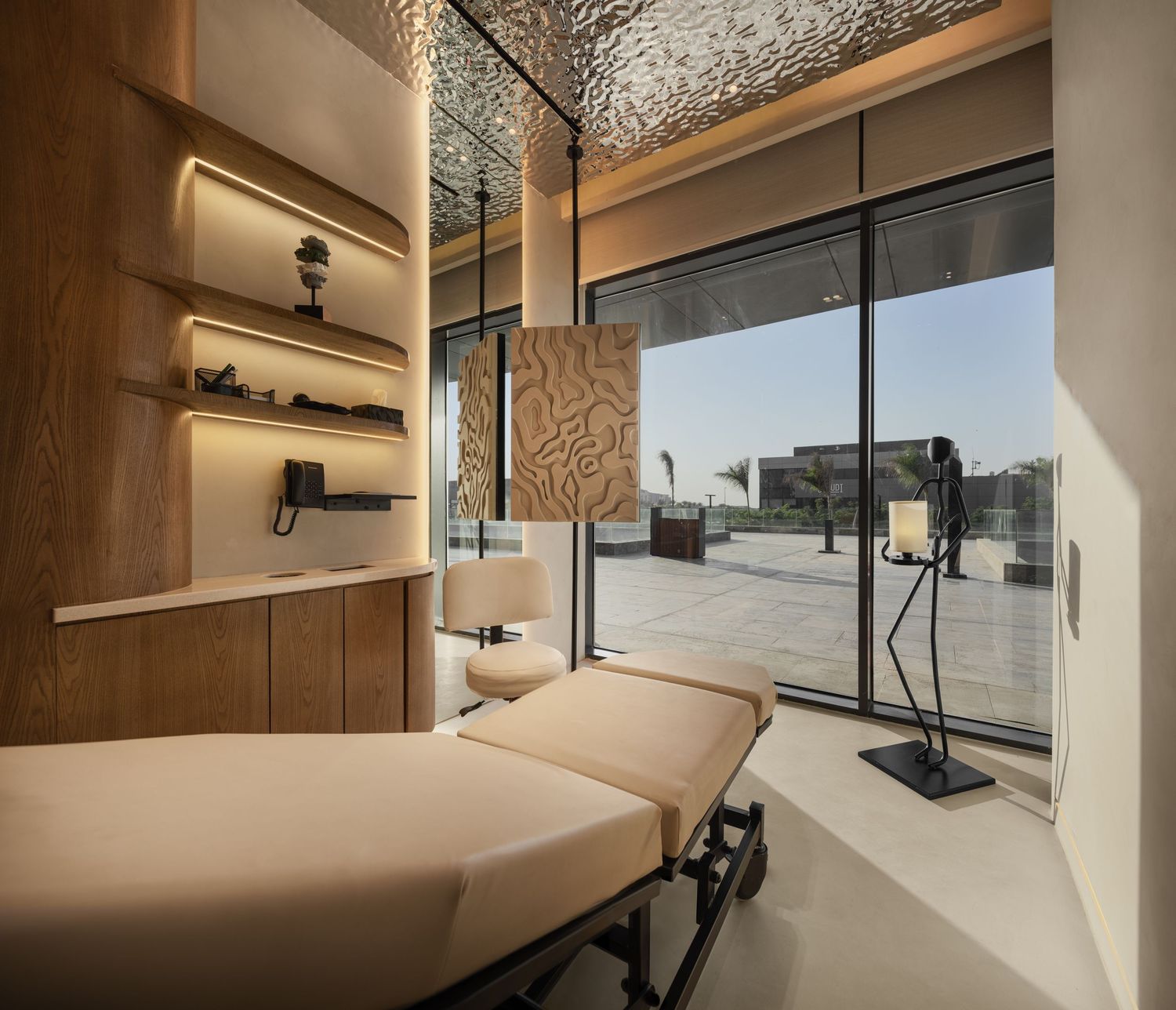 Luxury spa treatment room with beige massage bed, wooden shelves, textured lamp, and panoramic glass windows.