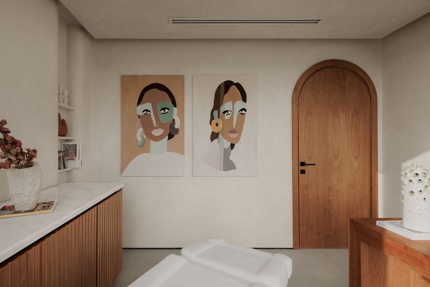 Serene spa treatment room with arched wooden door, abstract face artwork, and a white treatment table.