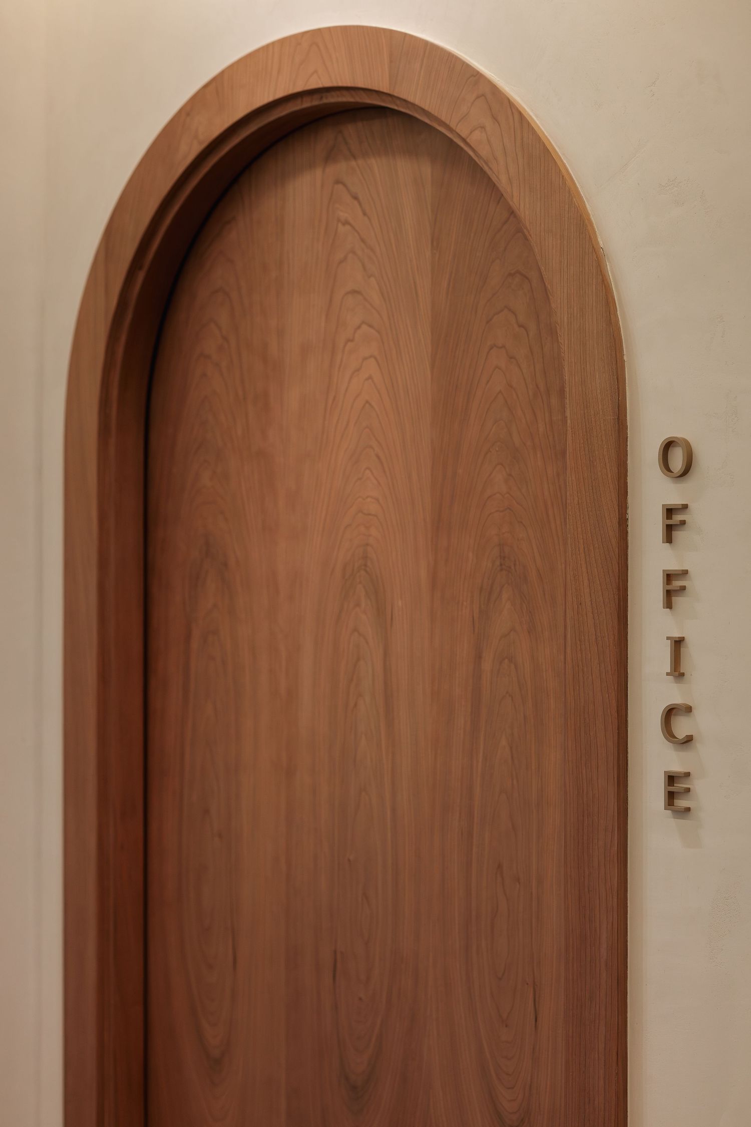 Elegant arched wooden door with natural wood grain texture beside vertical OFFICE letter signage on cream wall.
