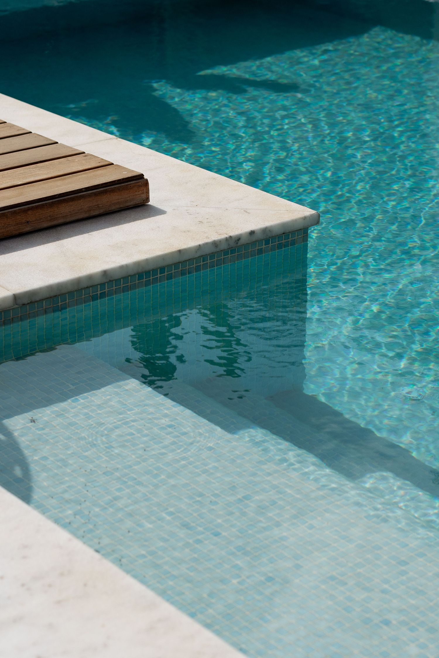 Detail of swimming pool with wooden deck, white marble edge, and turquoise mosaic tiles.
