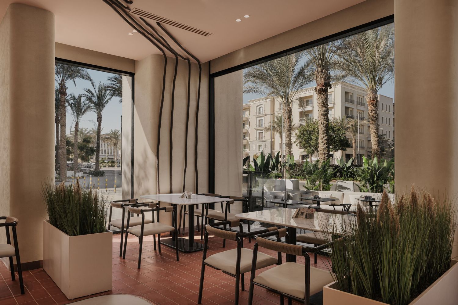 Bright restaurant dining area with large windows overlooking palm trees, featuring minimalist tables and chairs with potted plants.