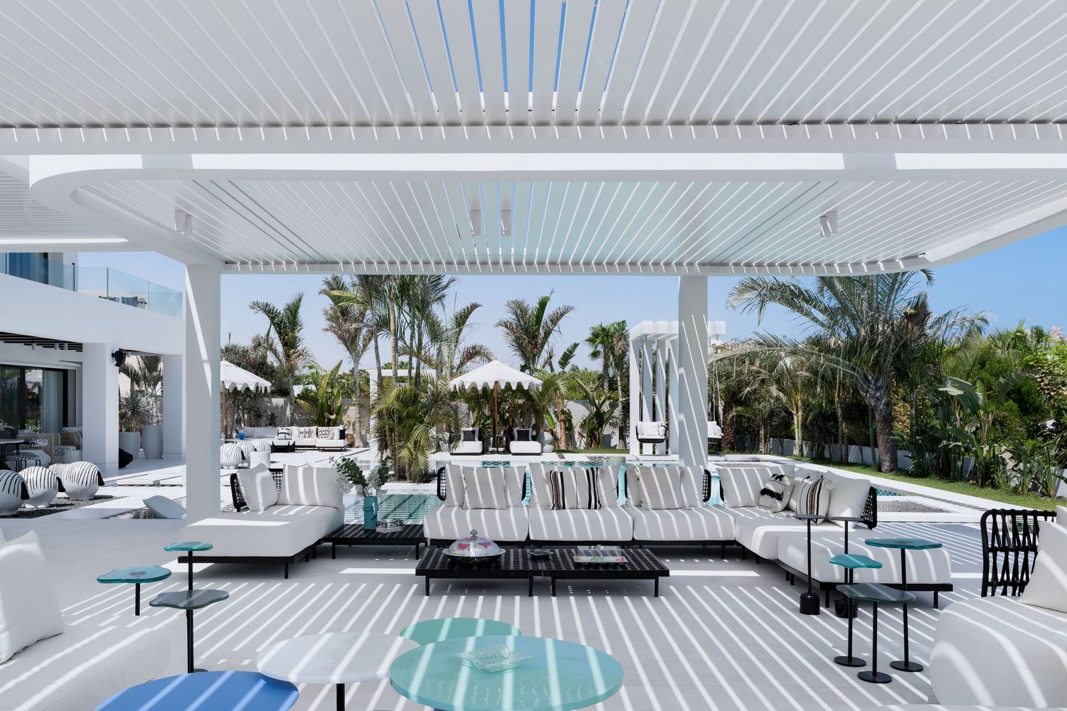 Spacious white outdoor living area with plush seating, teal rug, and palm trees under an open pergola.