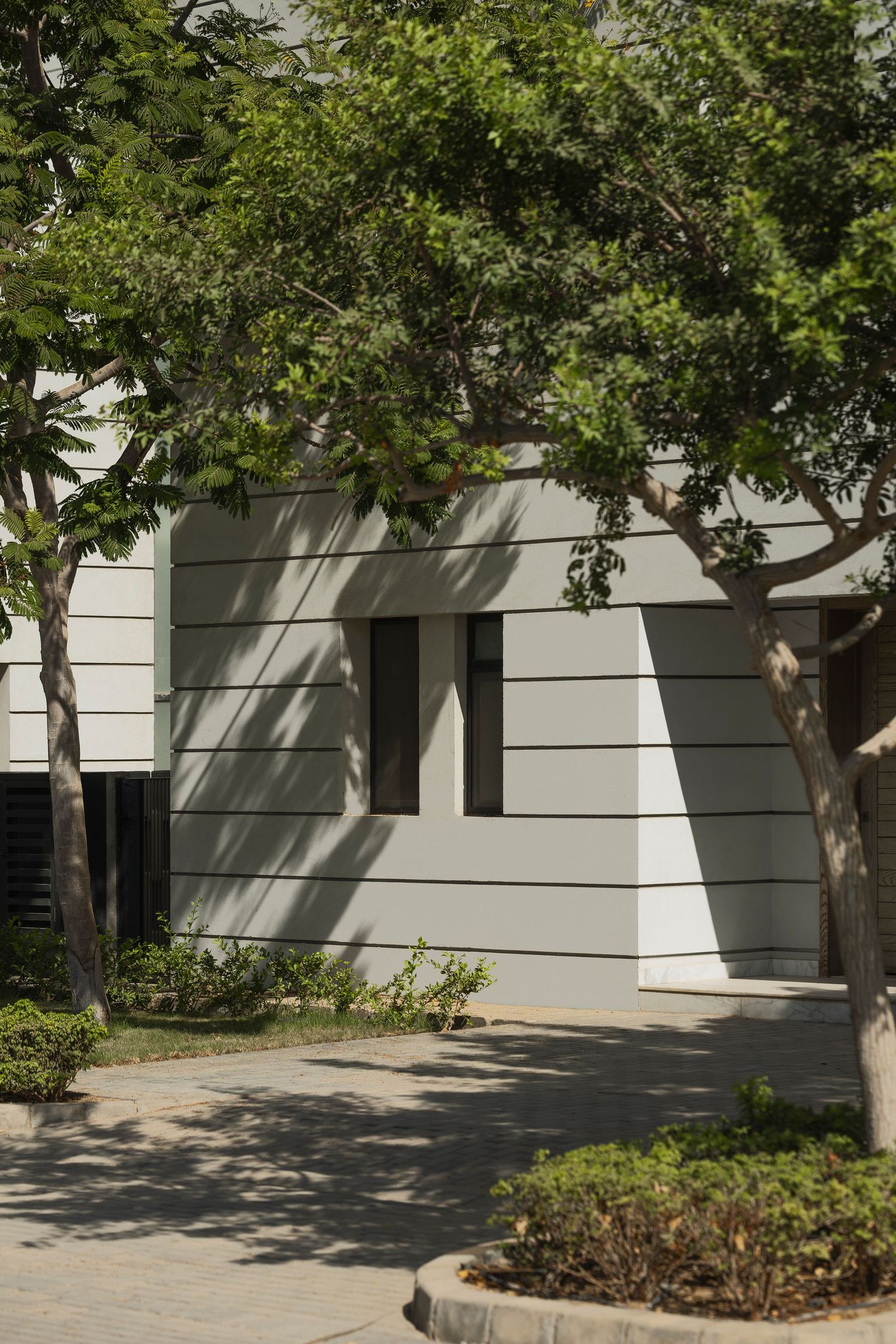 Modern white building facade with horizontal lines, framed by lush green trees casting shadows on a sunny day.