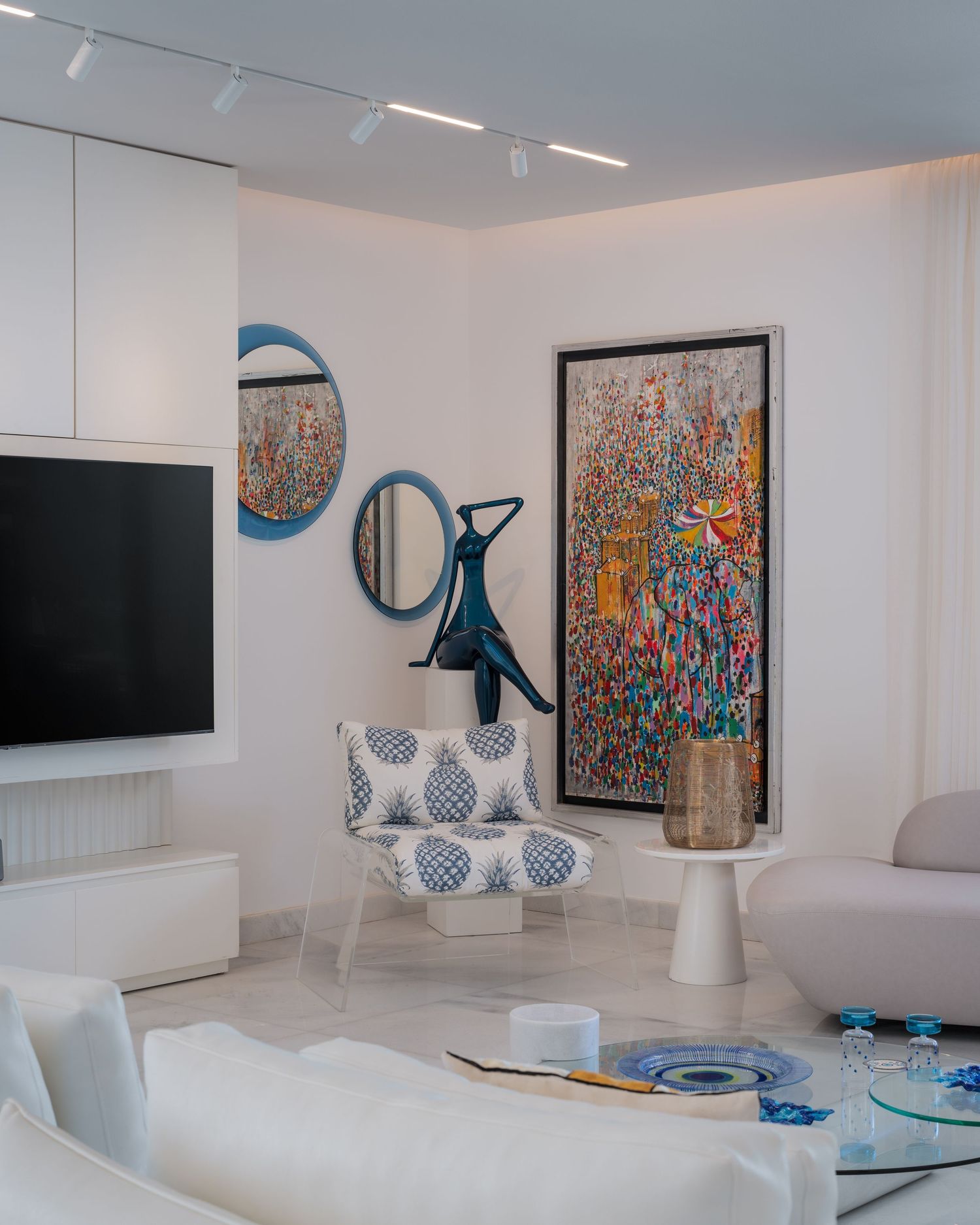 Bright white living room with colorful large artwork, blue round mirrors, sculptural decor, and patterned accent chair.