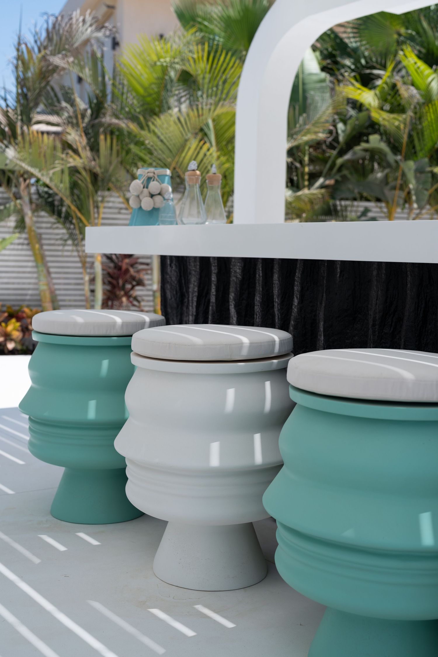 Three sculptural teal and white bar stools at an outdoor white bar counter with tropical palm tree backdrop.