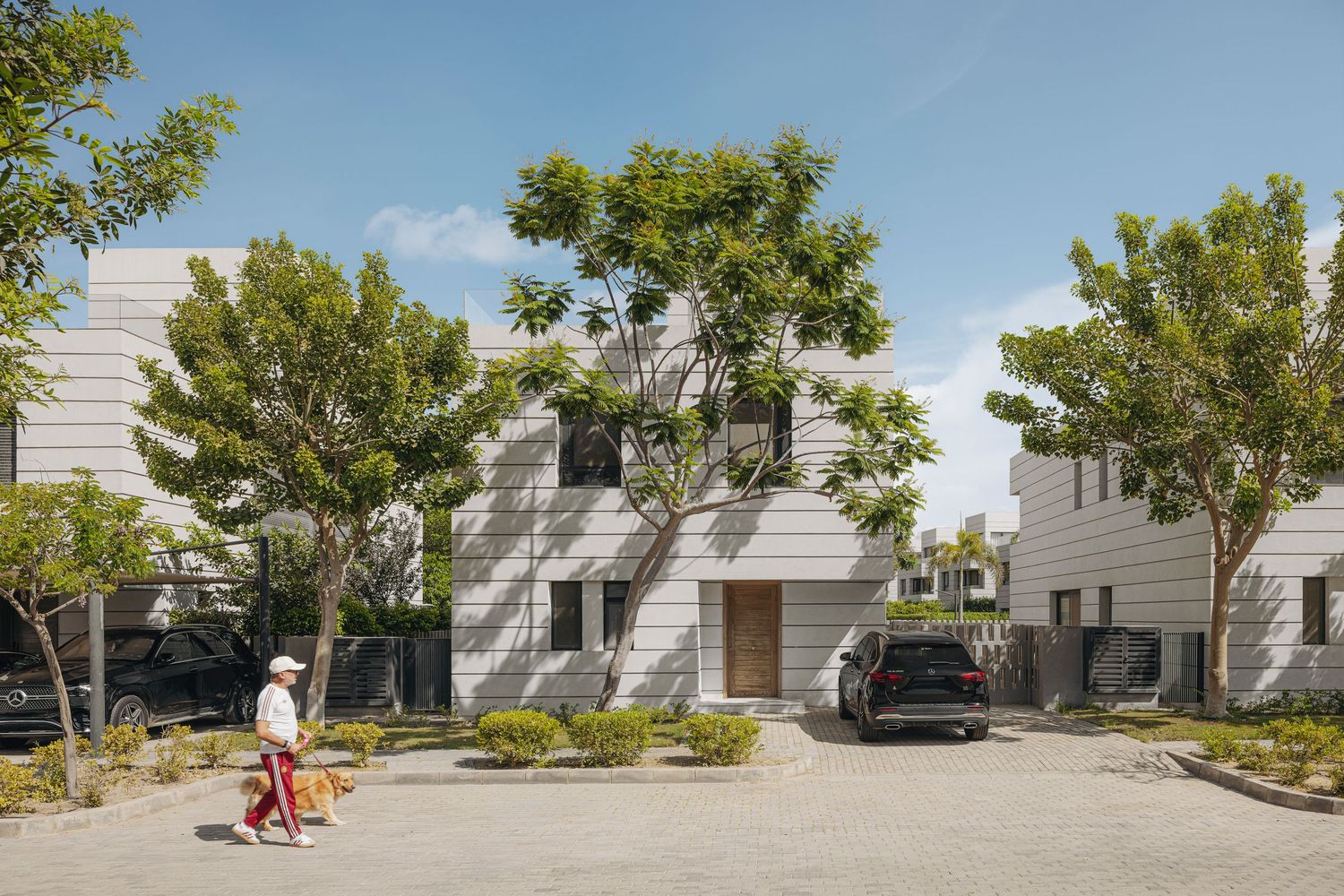 Modern white residential buildings with mature trees, landscaped grounds, and a pedestrian walking past parked cars on a sunny day.