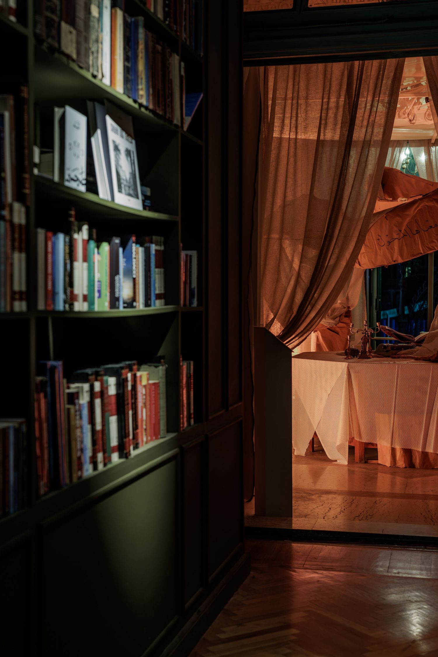Dark green bookshelf filled with books beside open doorway with flowing terracotta curtains and warm light.