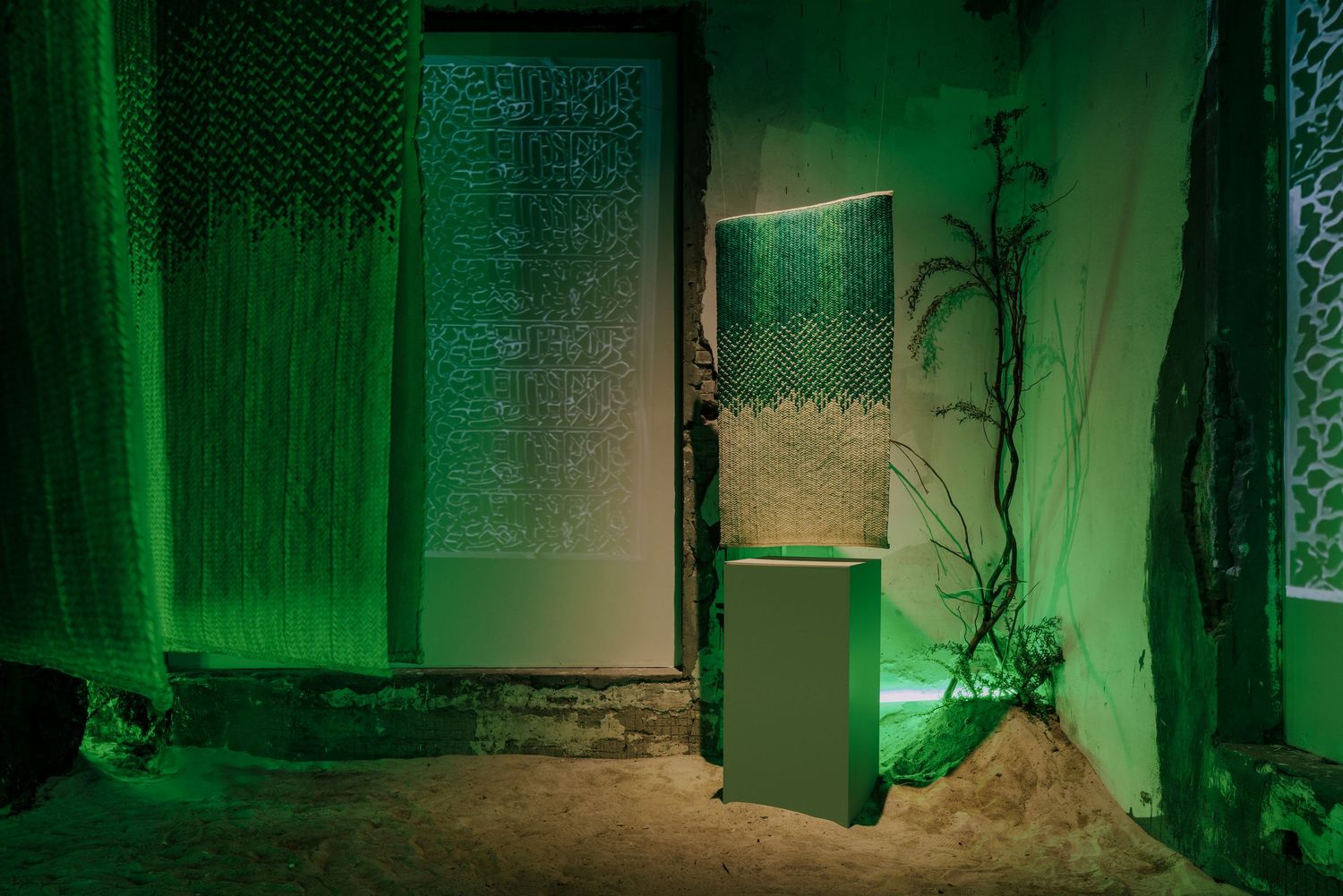 Dramatic green-lit installation with textured screens, tropical plants, and a white pedestal on sandy ground.