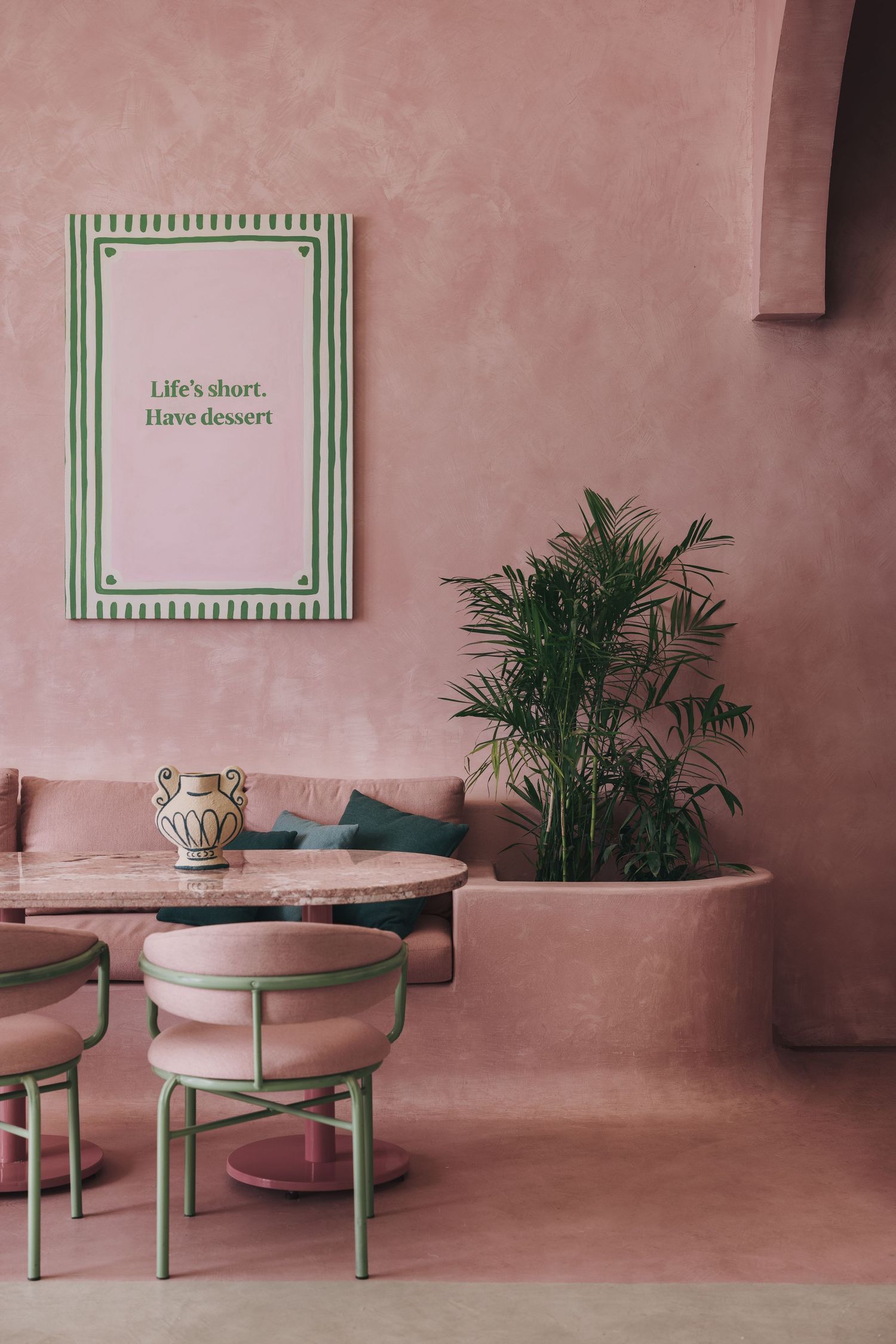 Pink cafe interior with green-framed motivational poster, marble table, palm plant and curved banquette.