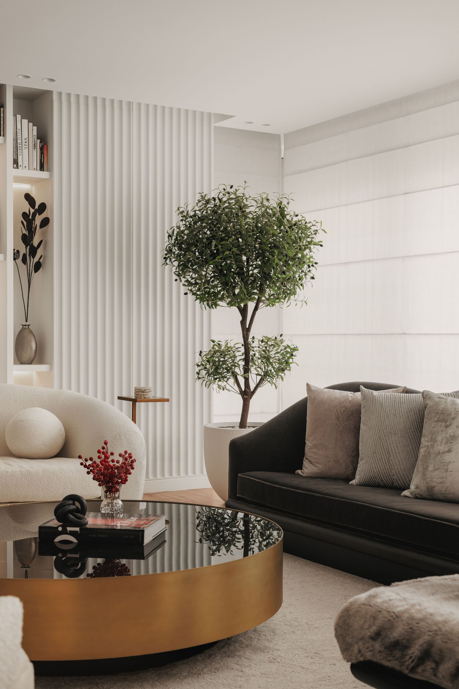 Contemporary living room featuring a dark sofa, white bouclé chair, gold glass coffee table, and large indoor olive tree.