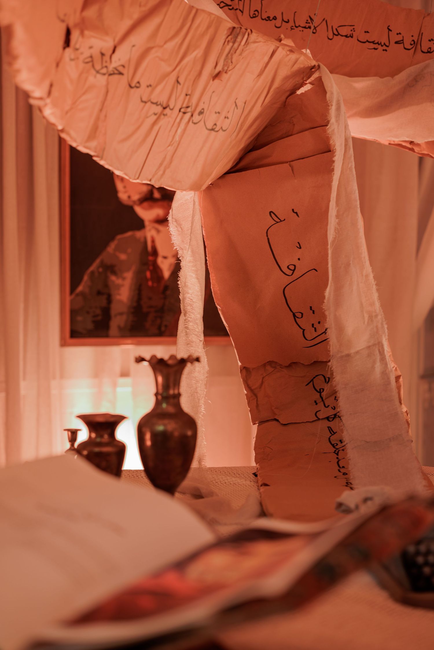 Hanging paper sheets with Arabic calligraphy script in a warmly lit artistic installation with decorative vases.