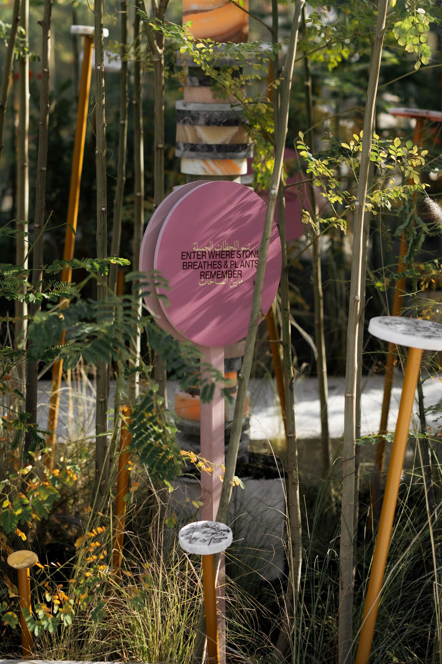 Garden art installation featuring a pink circular sign reading Enter Where Stone Breathes and Plants Remember.