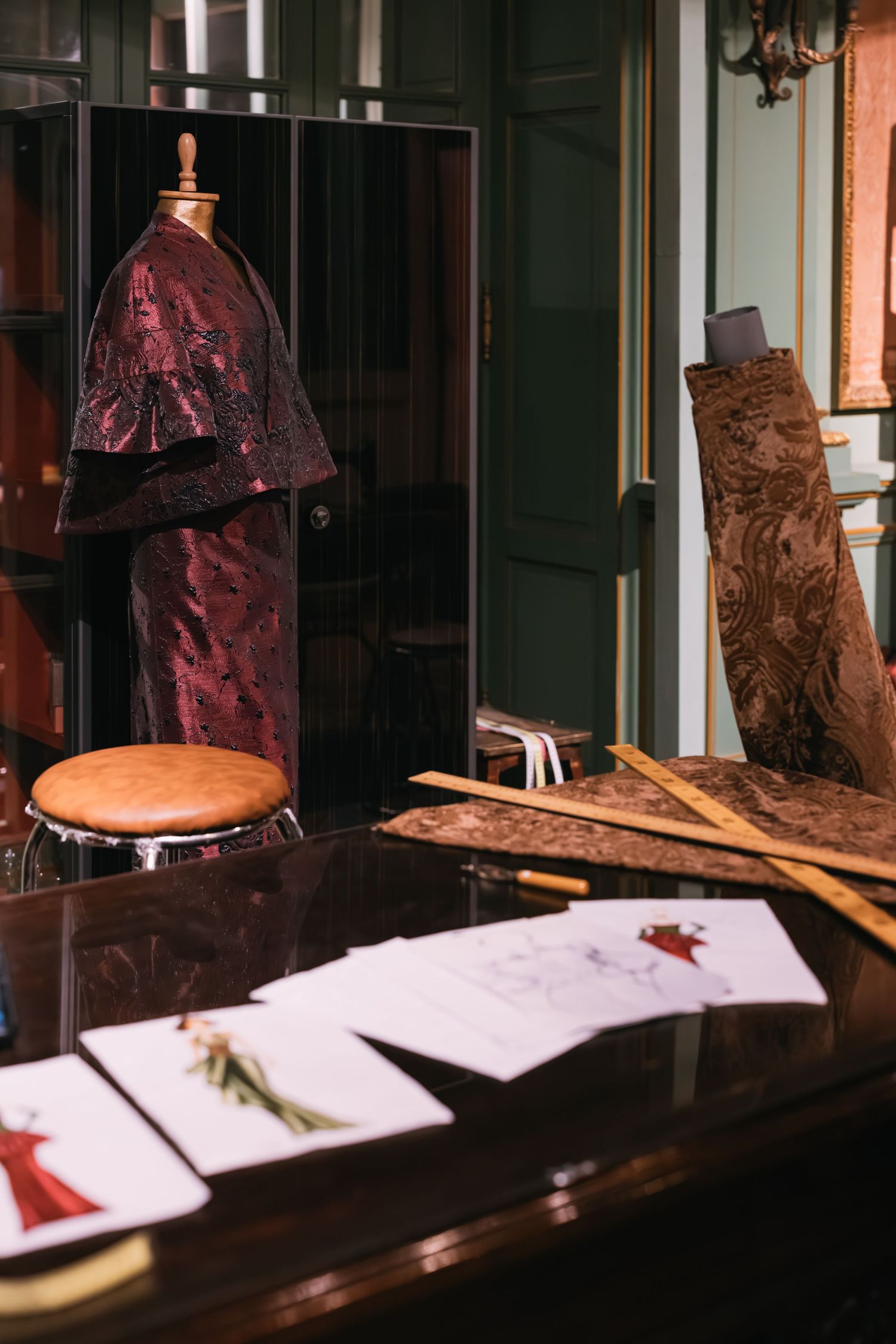 Burgundy brocade gown on mannequin in elegant dark green fashion atelier with design sketches on desk.