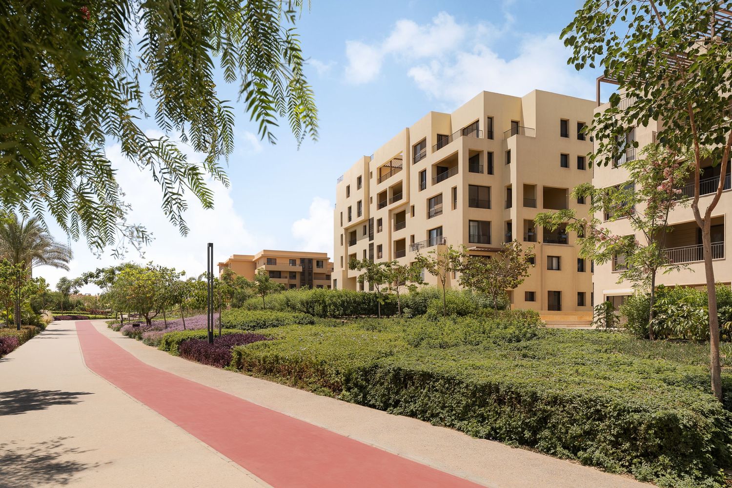 Sandy-toned apartment buildings with lush green gardens and red pathway lined with palm trees.