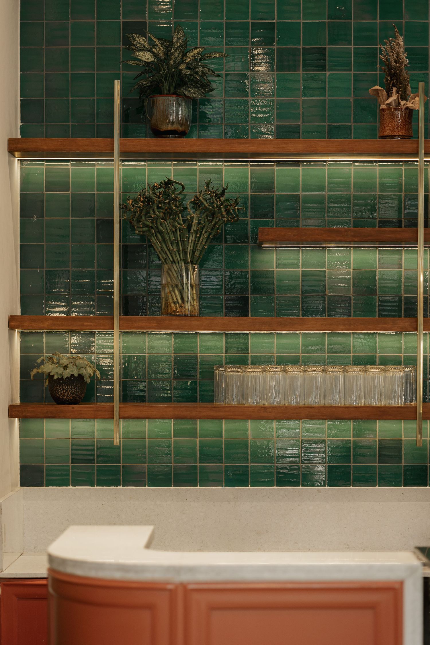 Green glazed tile wall with wooden shelves displaying potted plants and glassware above a terracotta cabinet.