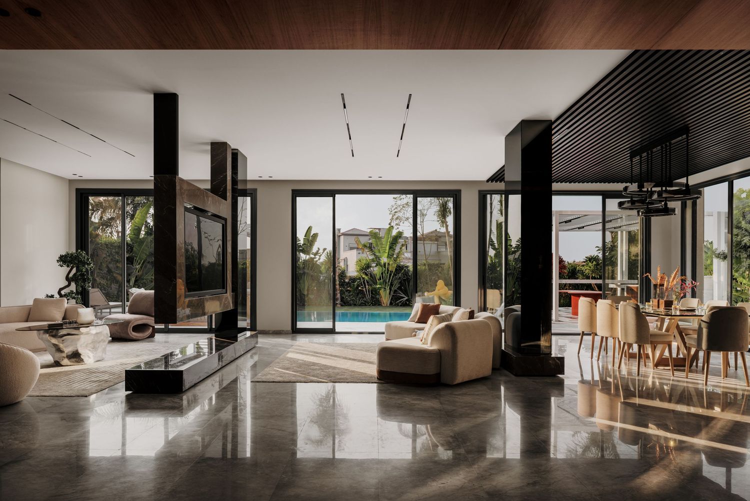 Spacious open-plan living area with dark columns, reflective floors, and floor-to-ceiling windows overlooking a pool.