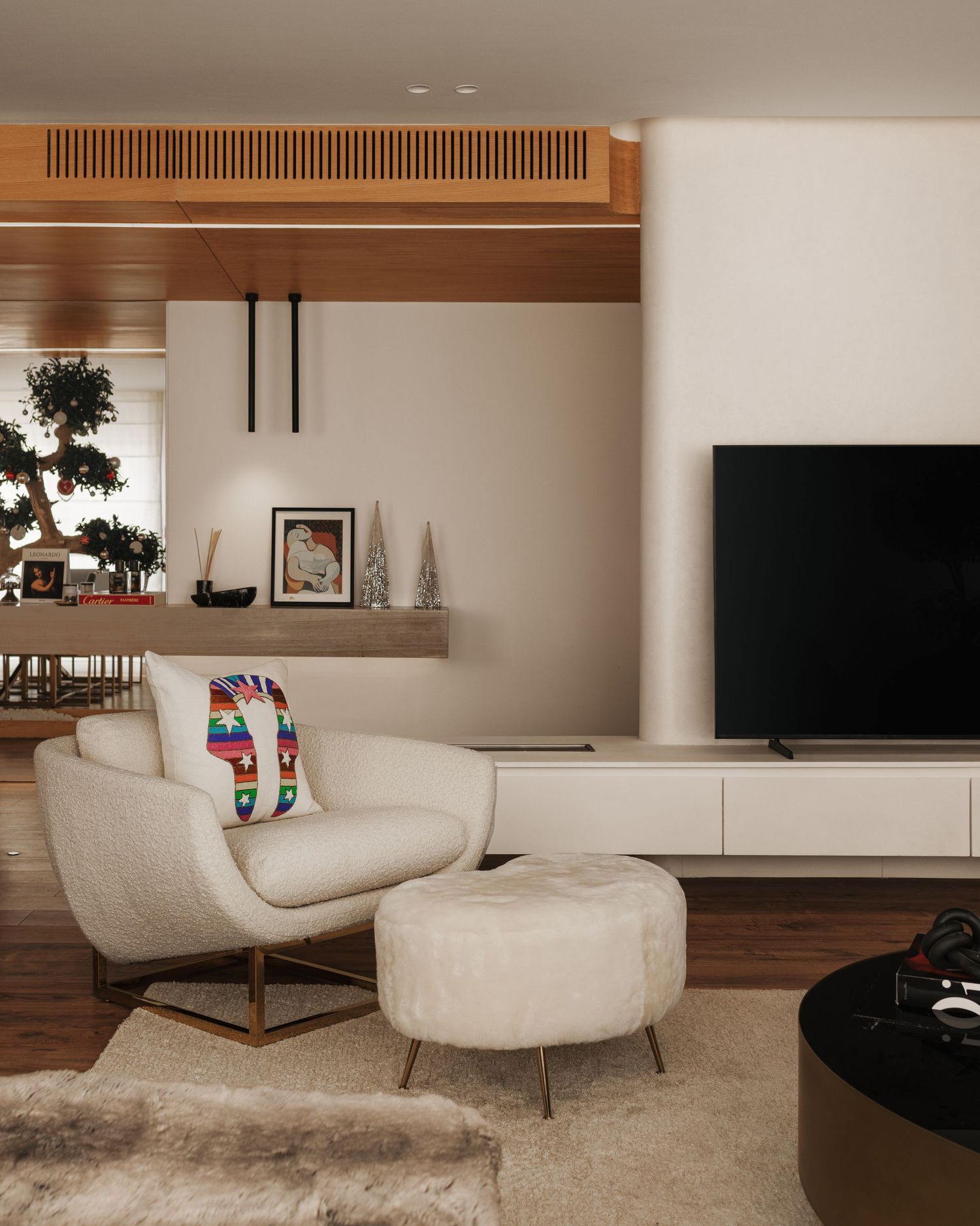 Contemporary living room with white bouclé armchair, ottoman, wall-mounted TV, wooden ceiling detail, and colorful accent pillow.