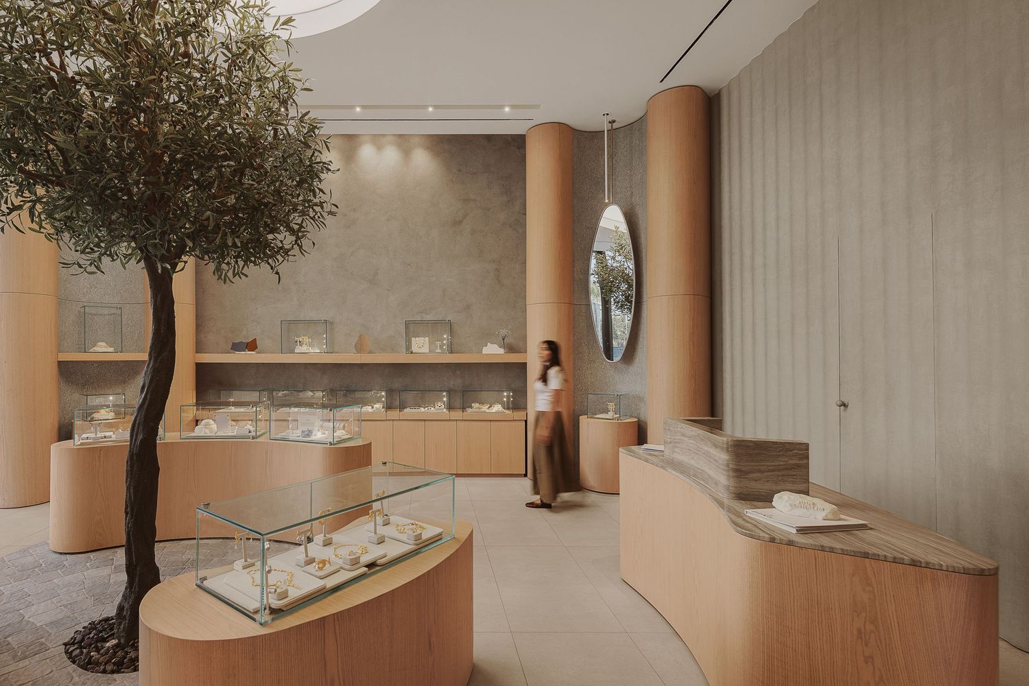 Spacious jewelry boutique with live olive tree, curved wooden displays, oval mirror, and a browsing customer.