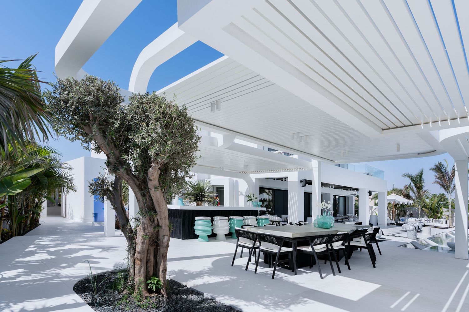 Modern white outdoor dining area with pergola, olive tree centerpiece, black dining set, and turquoise accents under clear blue sky.