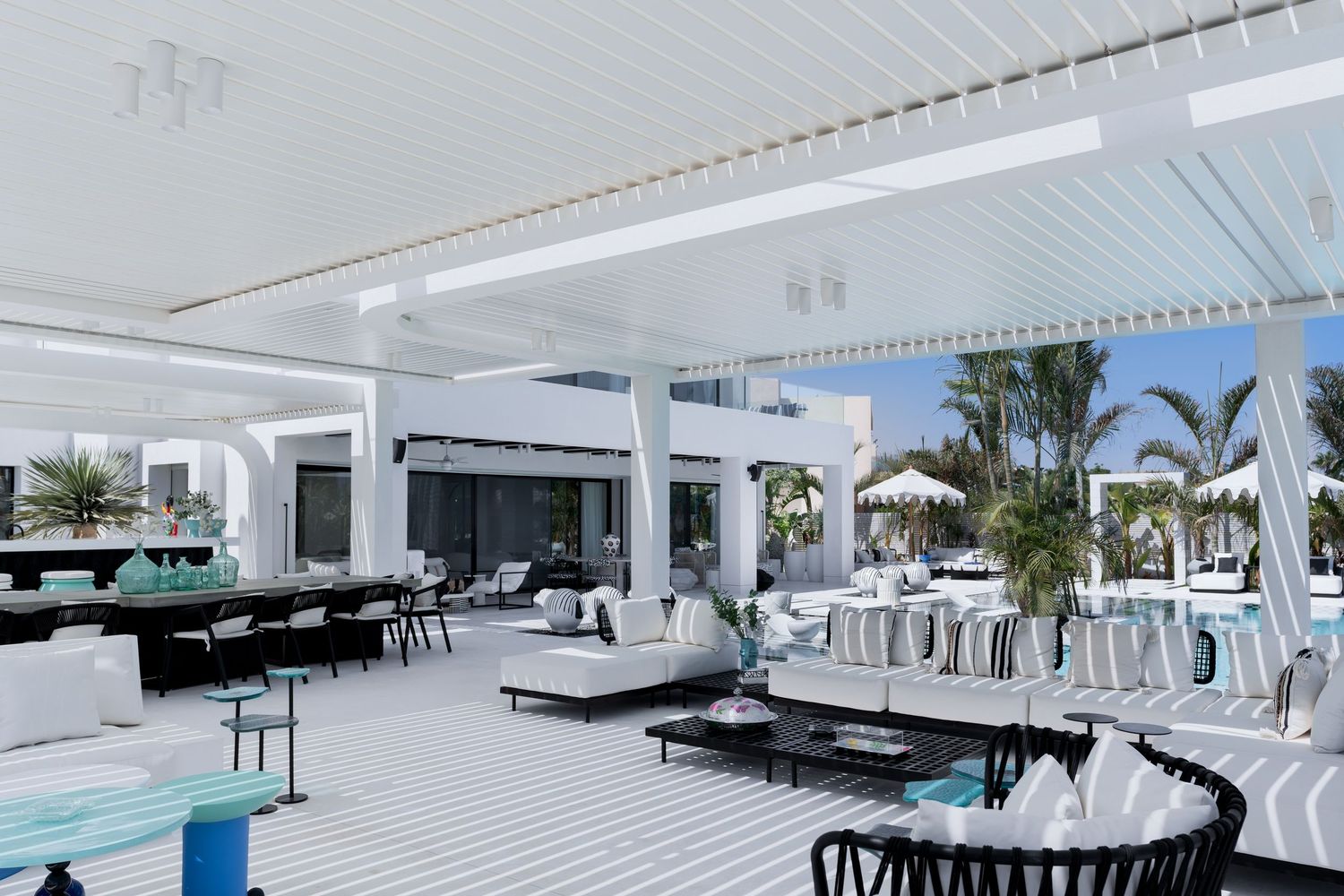 Luxurious white outdoor lounge area with striped flooring, black accents, and tropical palm trees under a pergola.