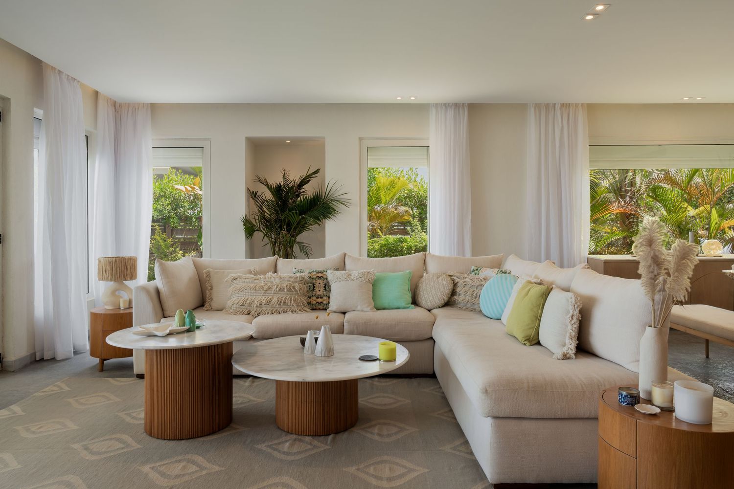 Bright luxury living room with L-shaped cream sofa, colorful cushions, round marble coffee tables, and lush tropical garden views.