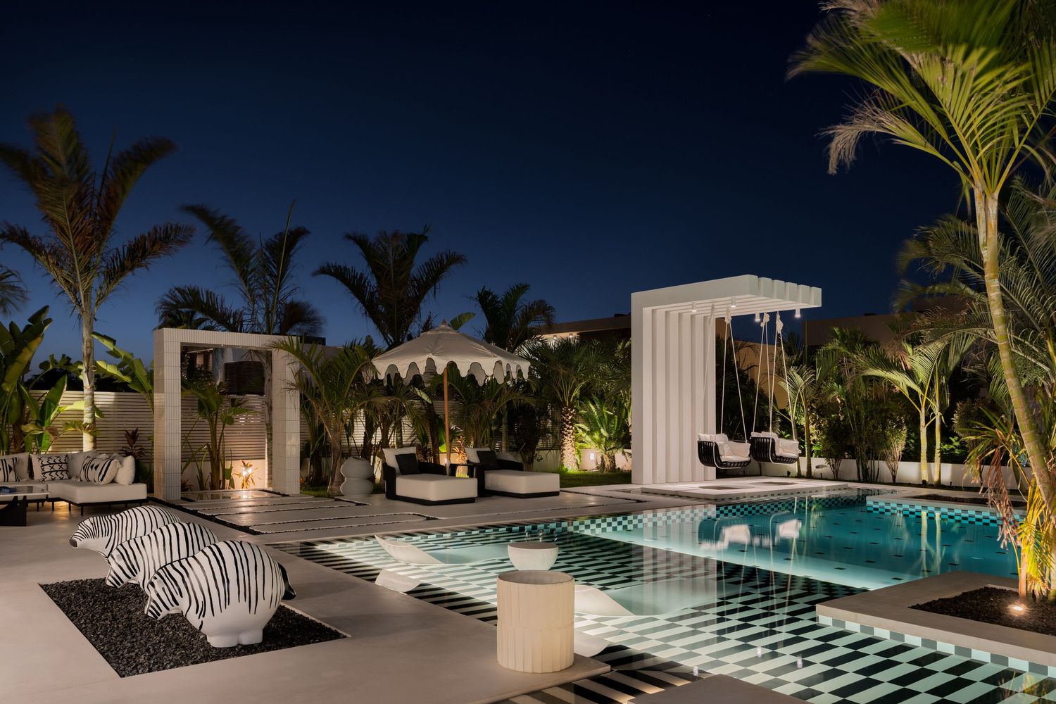 Exotic luxury pool area at night with checkered tiles, white cabanas, tropical palms, and decorative zebra-print sculpture.