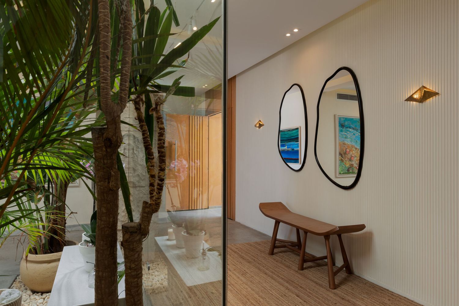 Elegant hallway with tropical plants, oval mirrors, wooden bench, and a serene bathroom visible through glass partition.