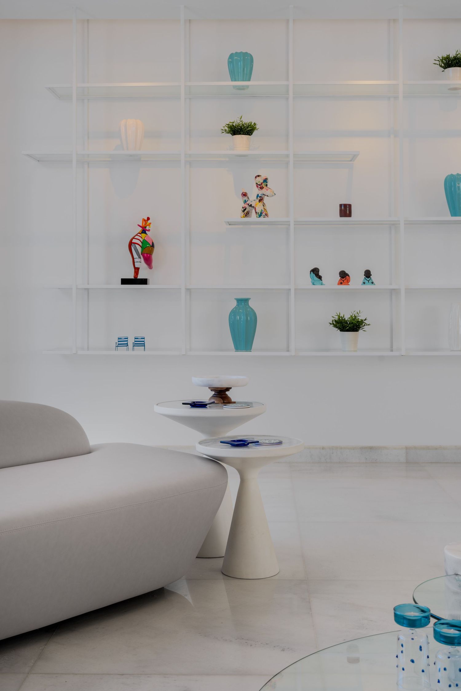 Minimalist white living room with open shelving displaying turquoise vases, colorful figurines, and a curved white sofa.