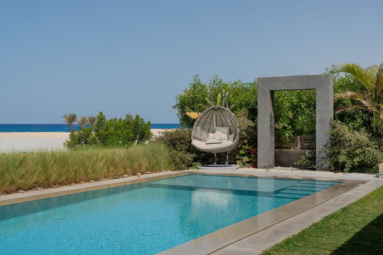 Private beachfront pool with hanging egg chair, stone archway, lush greenery, and direct sandy beach ocean access.