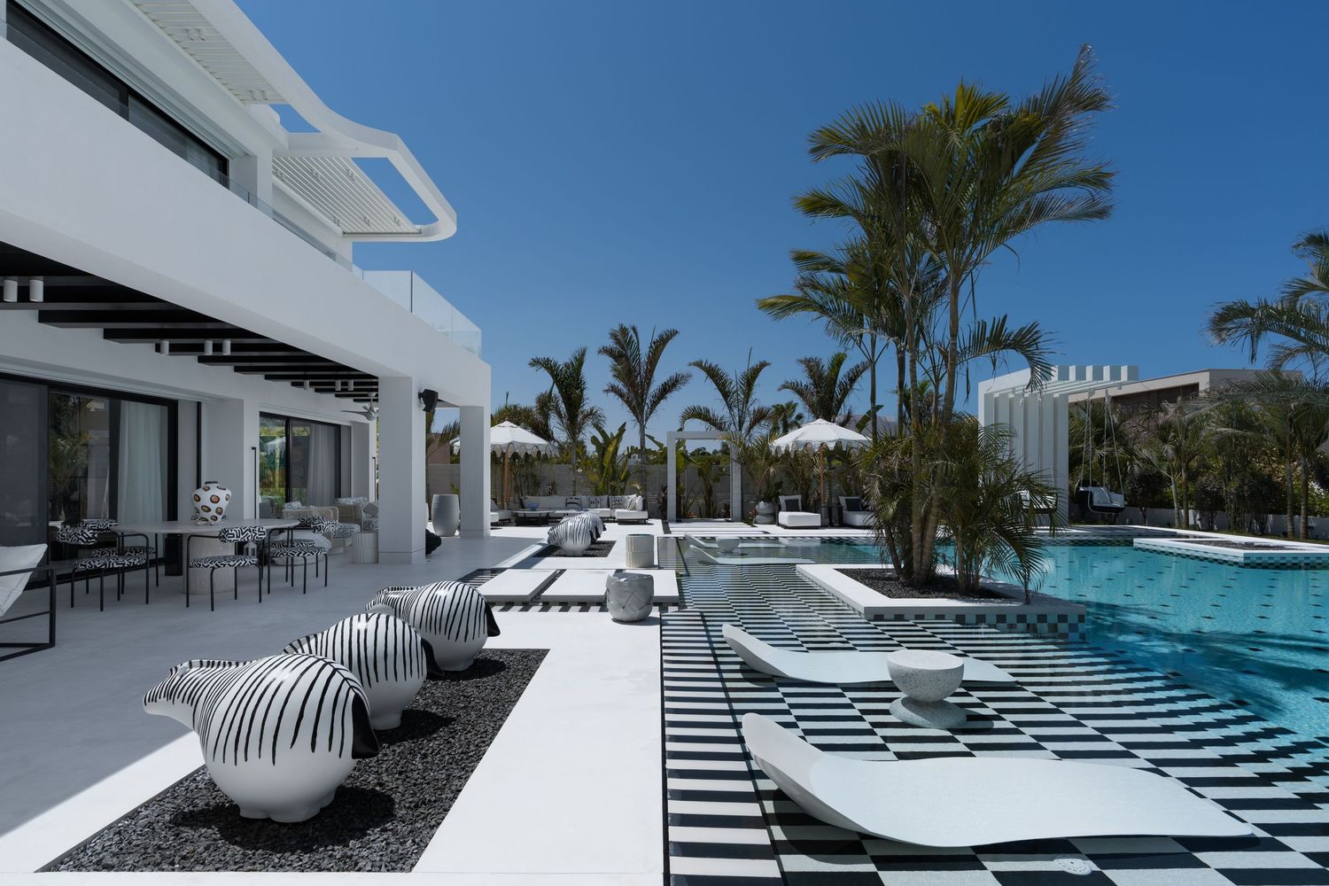Elegant white luxury villa with pool, black and white checkered tiles, silver sculptural decor, and palm trees.