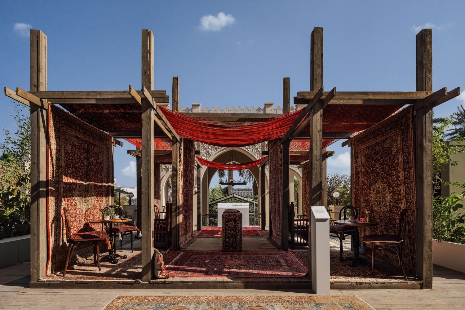 Rustic outdoor pergola with red draped fabric canopy and colorful traditional rugs on display under blue sky.