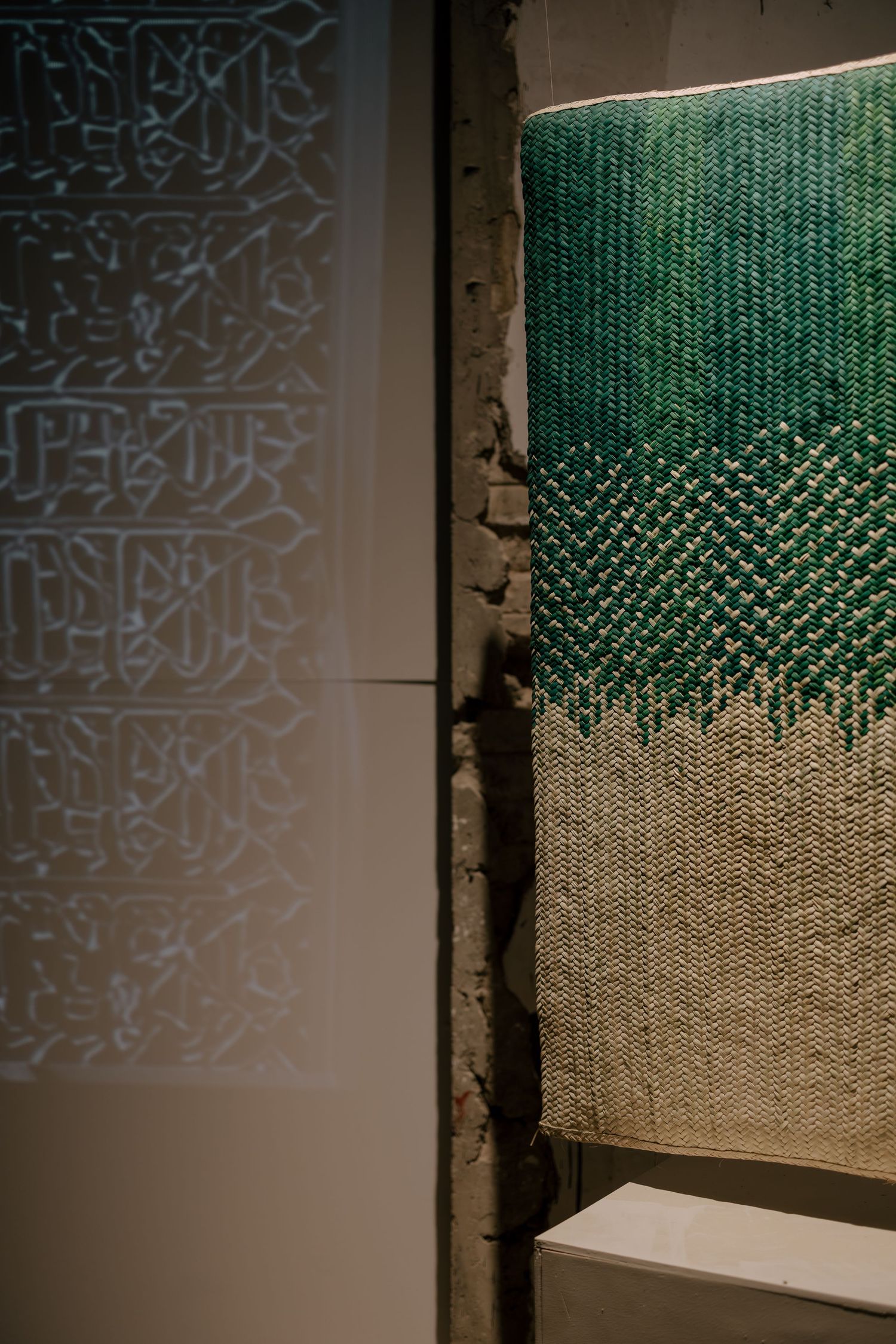 Green woven textile artwork beside illuminated Arabic calligraphy projection on crumbling white wall in dark gallery.
